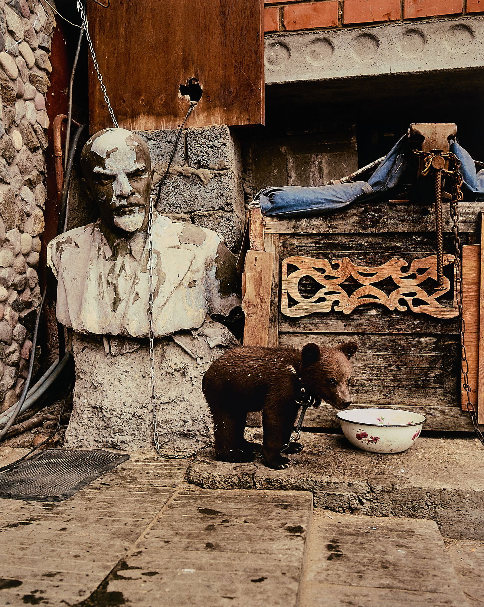 Moore "Misha & Vladimir, Lake Baikal" Photo 2003: Andrew Moore (American, b. 1957). C-Print photograph titled "Misha & Vladimir, Lake Baikal" depicting a toy bear chained beside a bust of Vladimir Lenin in disrepair, gazing outward amid a cluttered