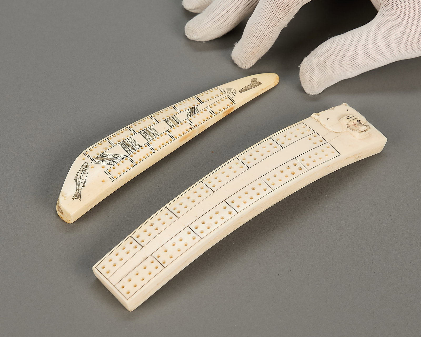 2 Inuit Cribbage Boards: Group of two Inuit carved organic material cribbage boards. One with black incised work depicting fish, seals and scale motifs. Unsigned, without pegs. One with carving of a polar bear's face and
