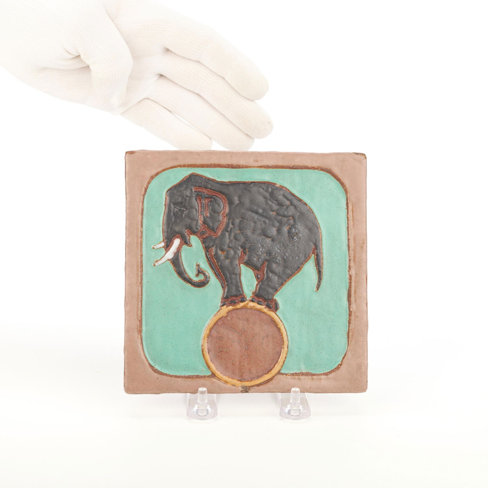 Mosaic Tile Co. Art Pottery Elephant Tile: Mosaic Tile Company, Zanesville, Ohio. American Arts & Crafts ceramic pottery tile depicting an elephant balancing atop a ball. With impressed mark along the verso.Height: 6 in x width: 6 in x depth: