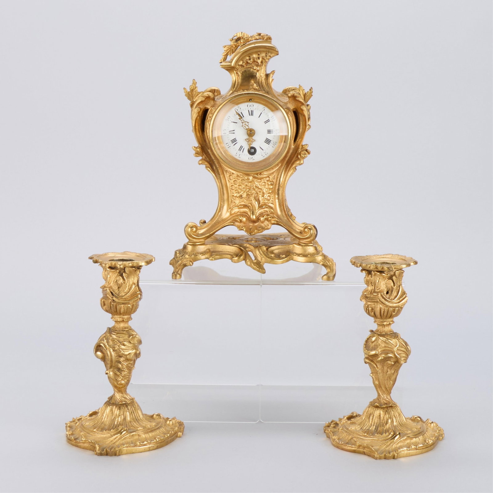 French Louis XV Style Ormolu Garniture Set: French Louis XV-style gilt bronze garniture set. Including a mantel clock and a pair of ormolu candlesticks, 19th-20th century. Decorated with Rococo scrollwork and foliate motifs. Provenance: The Dis