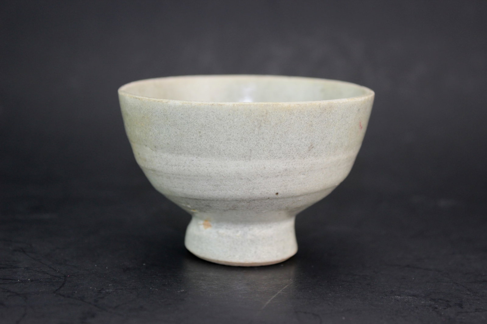 Old Chinese White Porcelain Bowl With Tall Foot: A small antique Chinese porcelain bowl featuring a subtle grayish glaze and a tall, rounded foot. The surface shows minor pitting, light glaze variations, and a few dark specks, consistent with age. S