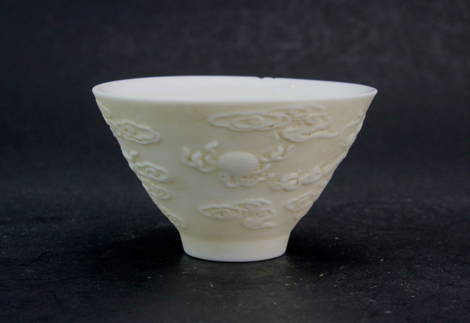 One Chinese White Porcelain Dragon Tea Cup: A white porcelain cup with carved dragon and cloud motifs, featuring a central sun design. It measures approximately 2 3/4 in. (H) X 1 1/2 in.(D). Chip on rim.