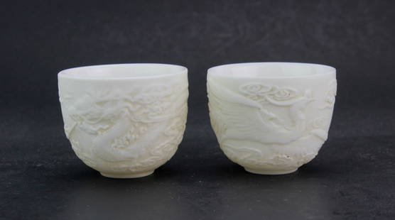 Pair of Chinese Blanc de Chine Porcelain Cups: A pair of Chinese blanc de chine porcelain cups featuring detailed reliefs of a dragon and a phoenix, symbolizing power and harmony, with cloud motifs and smooth glazed interiors. Each teacup measures