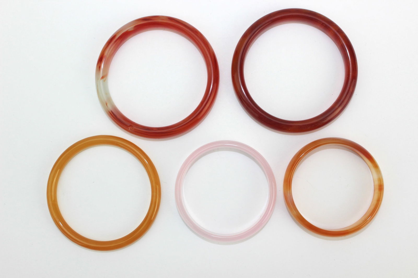 Group of Five Hard Stone Bangles: A group of five translucent hardstone bangles in red, orange, pink, and mixed hues. Minor surface wear on one, overall in good condition. Sizes range from approx. 67.7 mm to 53.1 mm (outer) and 54.7 m