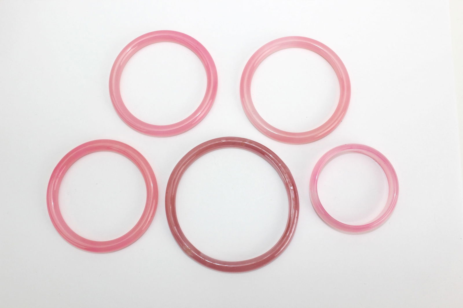 Set of Five Pink Hardstone Bangles: A group of five translucent pink hardstone bangles ranging from light rose to deep tones, with smooth surfaces, minor inclusions, and light wear. Largest approx. 77.7 mm (outer) × 64.7 mm (inner) dia