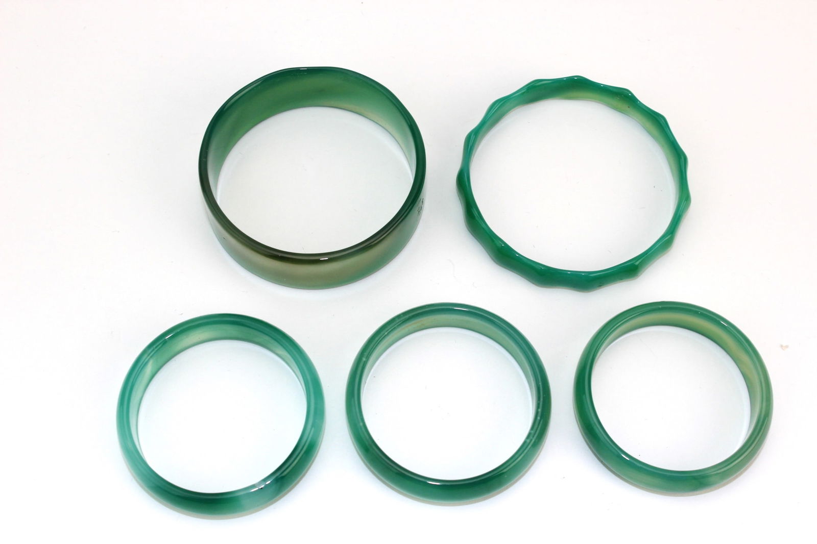 Lot of Five Green Hardstone Bangles: A group of five green hardstone bangles in varied shades and styles, including a wide band and a notched-edge piece, all smoothly polished with natural color variations. Minor inclusions, overall in g