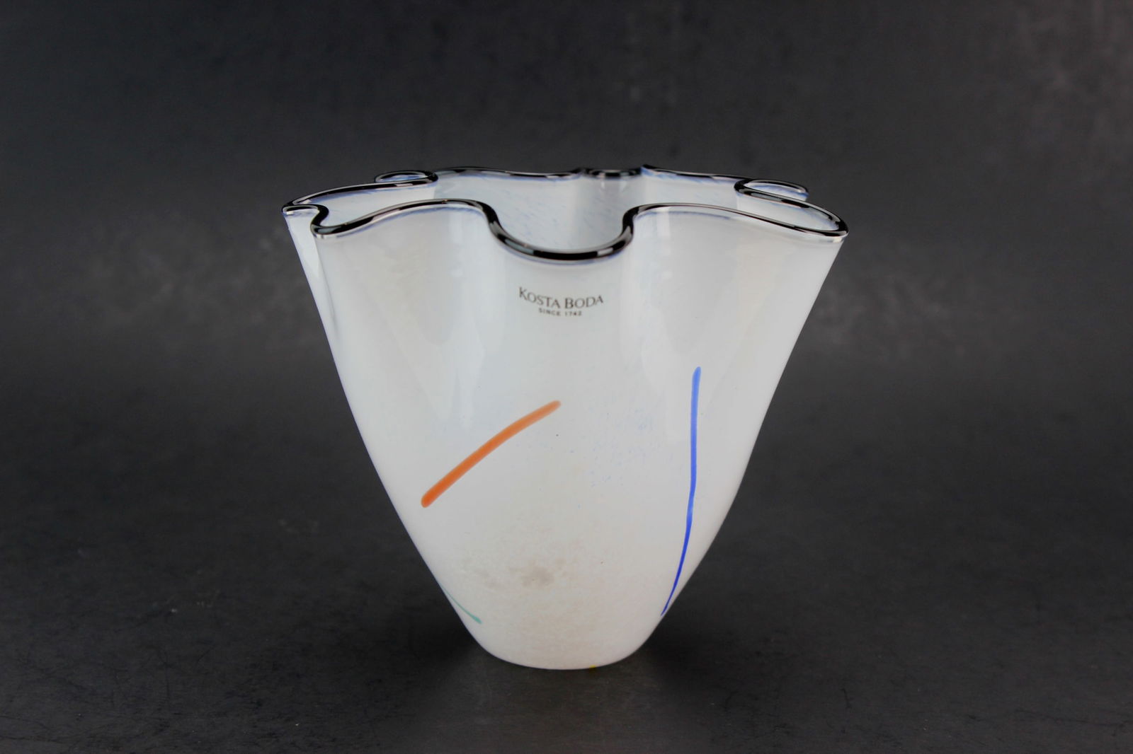 Kosta Boda Studio Art Glass Bowl by Ulrica HV: Kosta Boda studio art glass bowl by Ulrica Hydman-Vallien, model no. 49947, featuring a freeform ruffled rim, white ground with blue speckles, multicolor abstract accents, and a black-edged lip. Signe