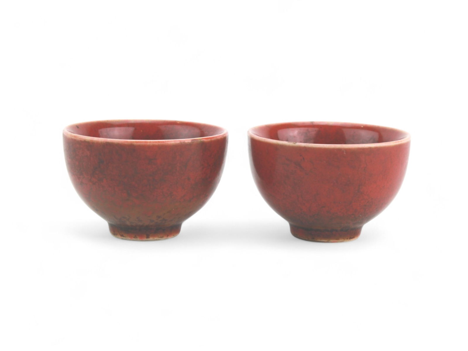 Two Chinese Oxblood Glazed Tea Cups: A pair of Chinese porcelain teacups featuring a deep copper red glaze with mottled tones, rounded bodies, and short unglazed foot rings. Each cup measures approximately 2 1/2 in. (D) X 1 3/4 in. (H).