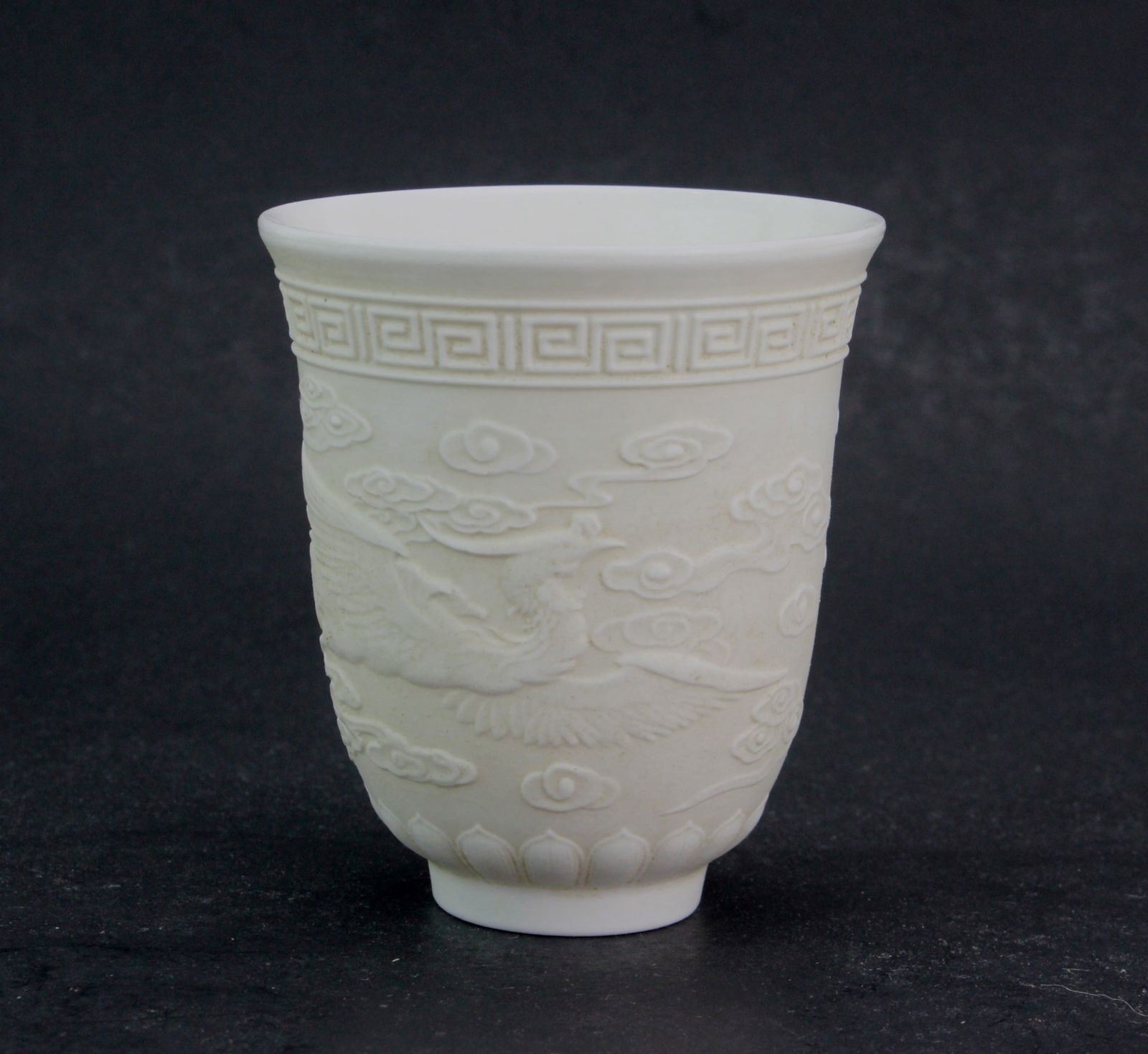 One Chinese White Porcelain Phenix Tea Cup: A white porcelain cup with a raised design of a phoenix among clouds, bordered by a meander pattern at the rim and lotus petals at the base. Unglazed exterior with a glazed interior. It measures appro
