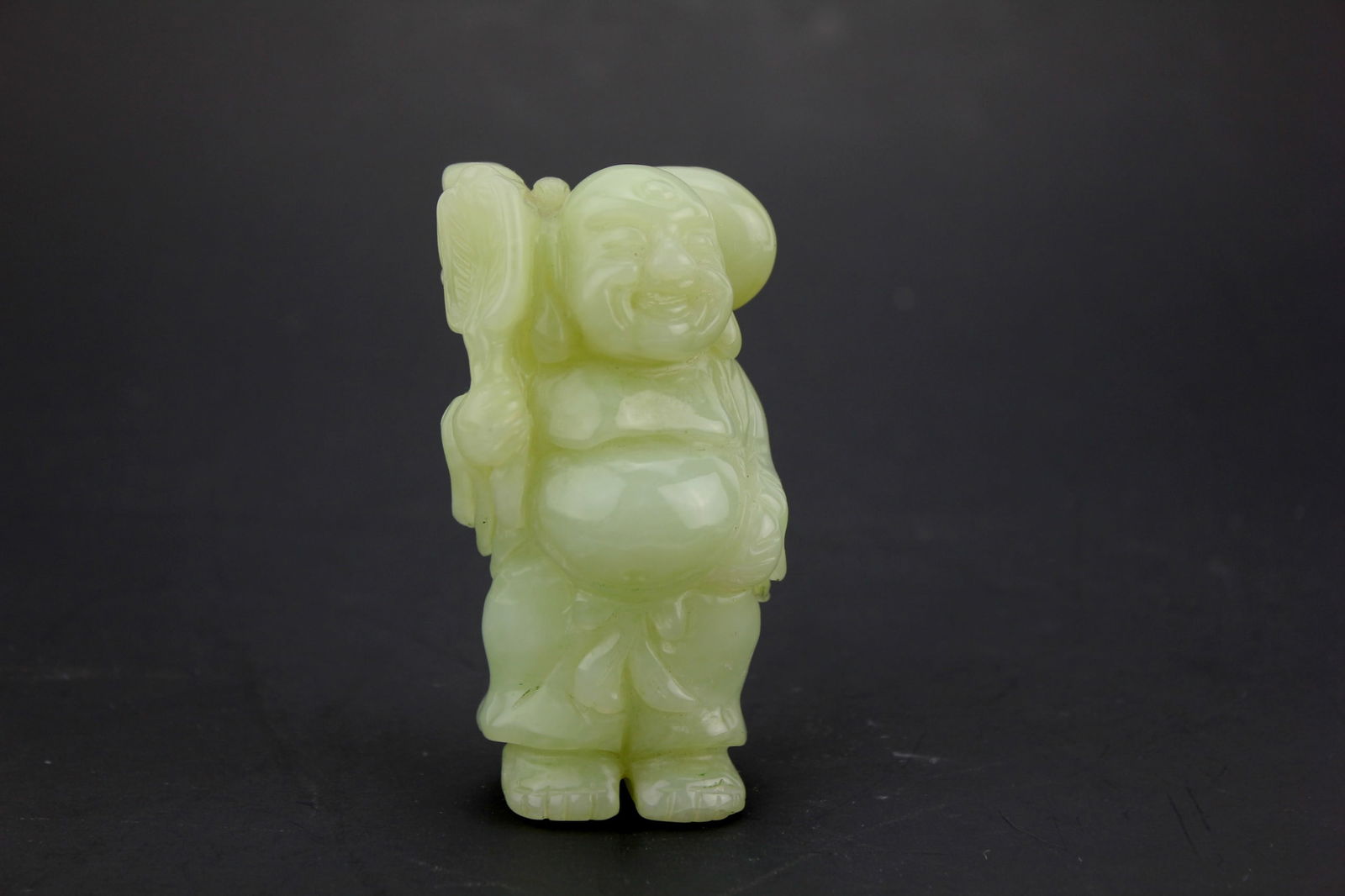 Chinese Jade Figurine of Laughing Buddha: A Chinese carved jade figurine standing 3 1/2 inches tall, depicting a joyful Buddha with a large pack over his shoulder. The stone has a smooth, translucent pale green tone. The carving is