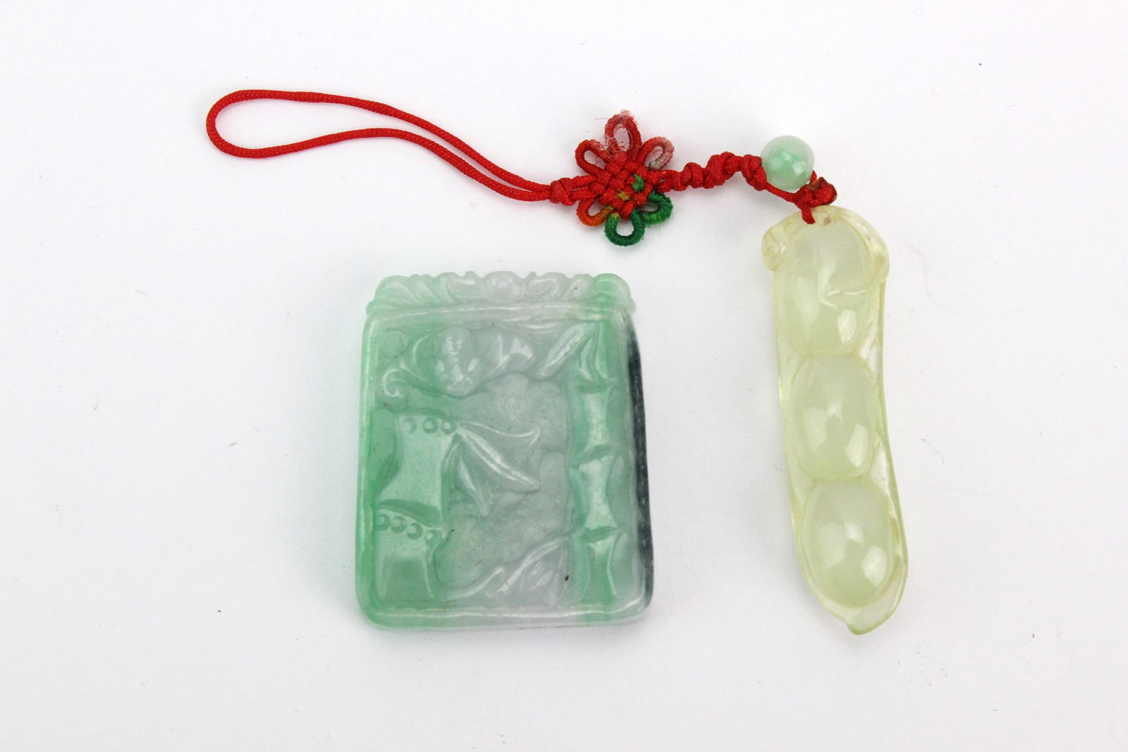Two Carved Jadeite Pendants: A pair of jadeite pendants: one square with floral and bamboo motifs in mottled green (approx. 34.7 × 28.6 mm), and one light green/yellow pea pod design with red cord (approx. 41 mm). Minor surface