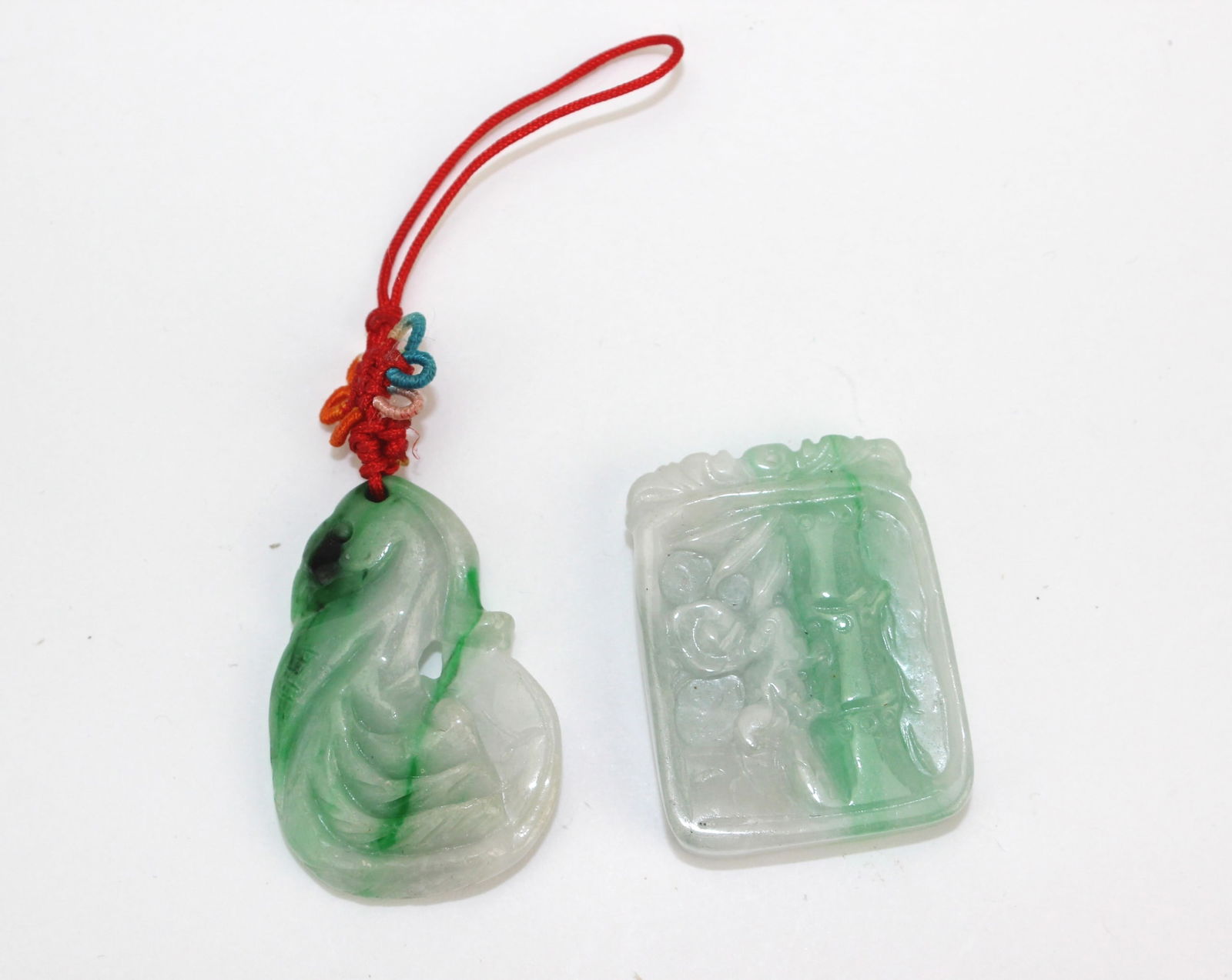 Two Carved Jadeite Pendants: A pair of jadeite pendants: one mottled green and white with naturalistic form on red cord (approx. 35.2 × 23.6 mm), and one rectangular pale green and white with floral and bamboo motifs (approx.
