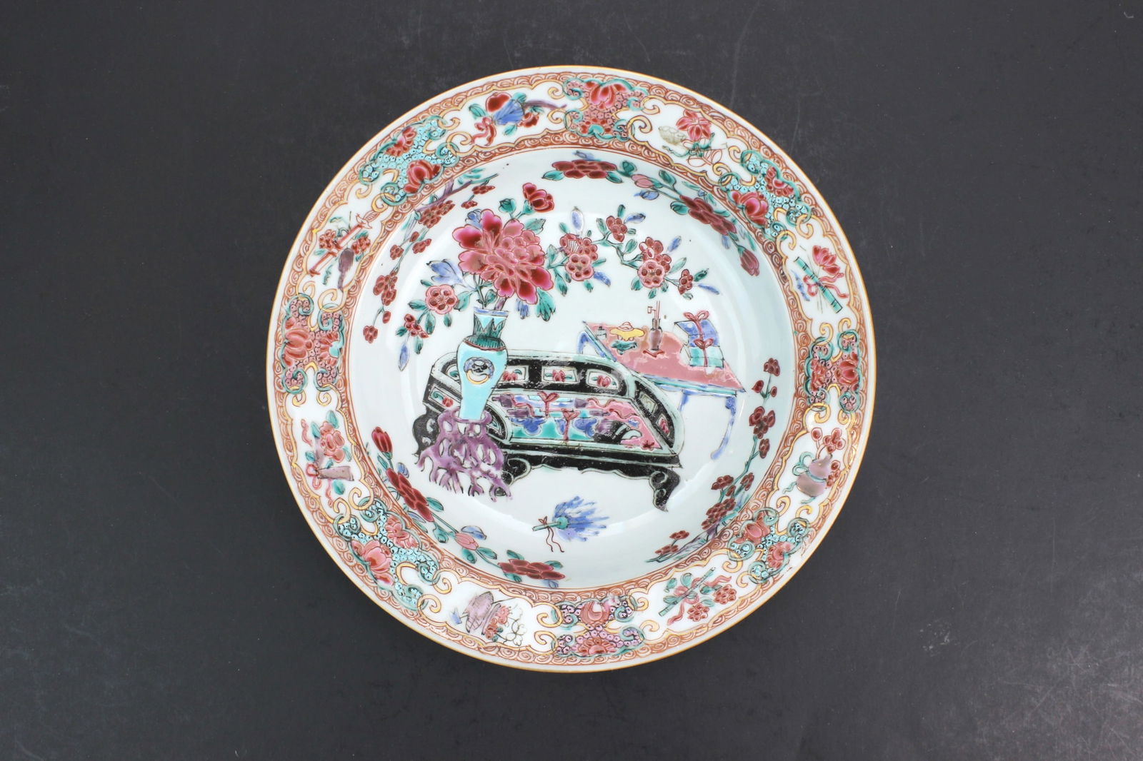 Chinese Export Famille Rose Deep Plate: Chinese Export porcelain deep plate finely decorated in famille rose enamels, featuring a central interior scene with flowers, scholar’s objects, and furniture, framed by a richly patterned floral