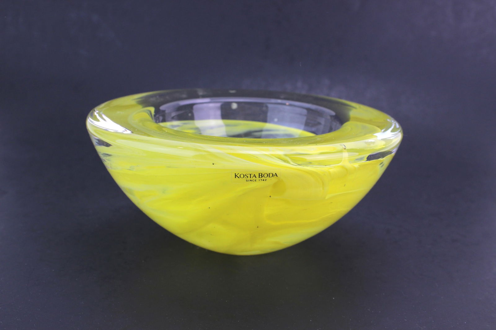 Kosta Boda Yellow Art Glass Bowl: Handcrafted Kosta Boda studio art glass bowl featuring a vibrant yellow swirl design with clear glass accents. Measures 6 3/4 in. (D) x 3 in. (H). In very good condition with no chips or cracks noted.