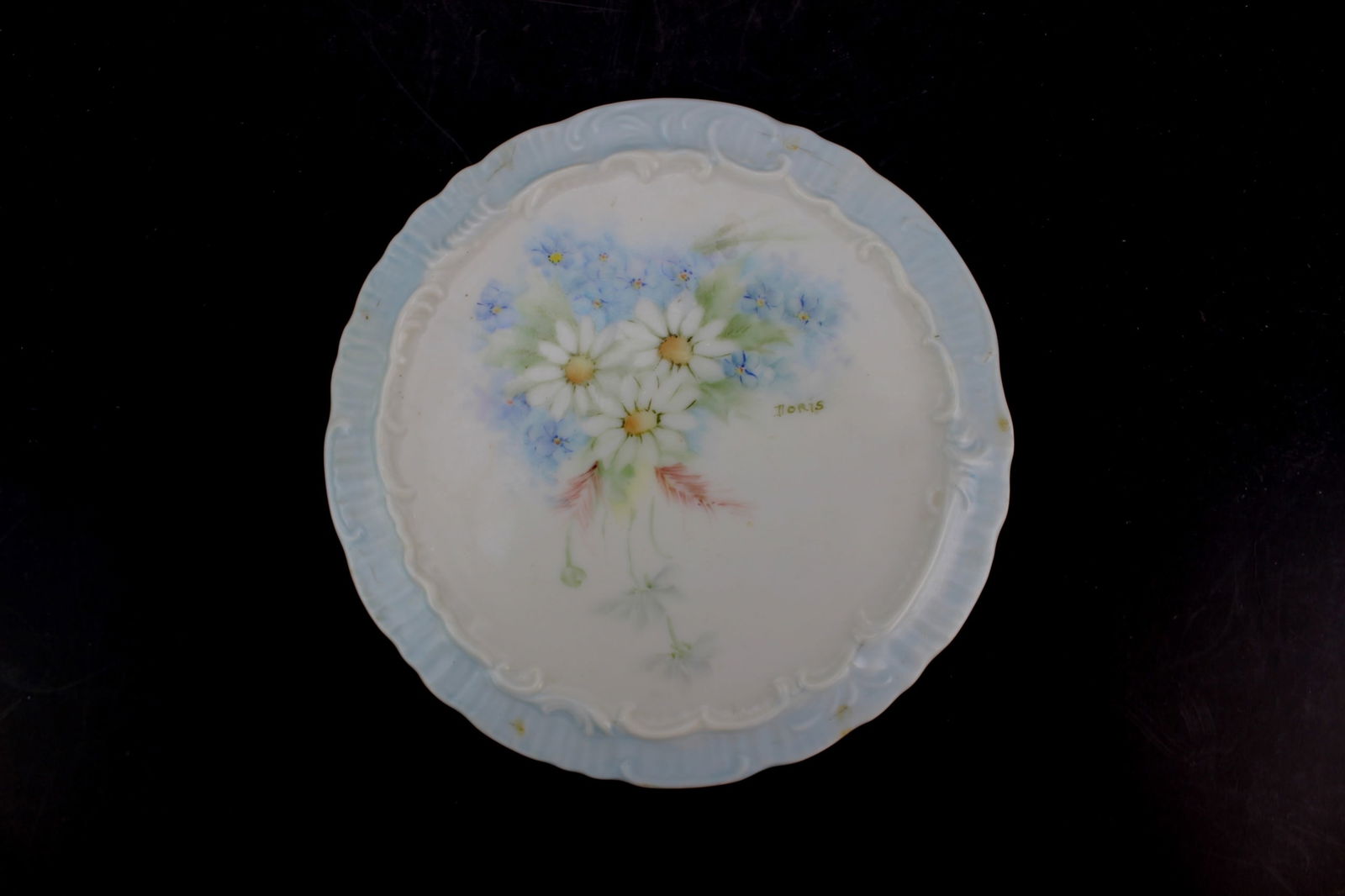 Hand-Painted Porcelain Trivet Signed by Loris: This decorative porcelain trivet features hand-painted white daisies and blue forget-me-nots, accented with soft greenery and signed "IORIS." The plate has a light blue scalloped rim and raised relief