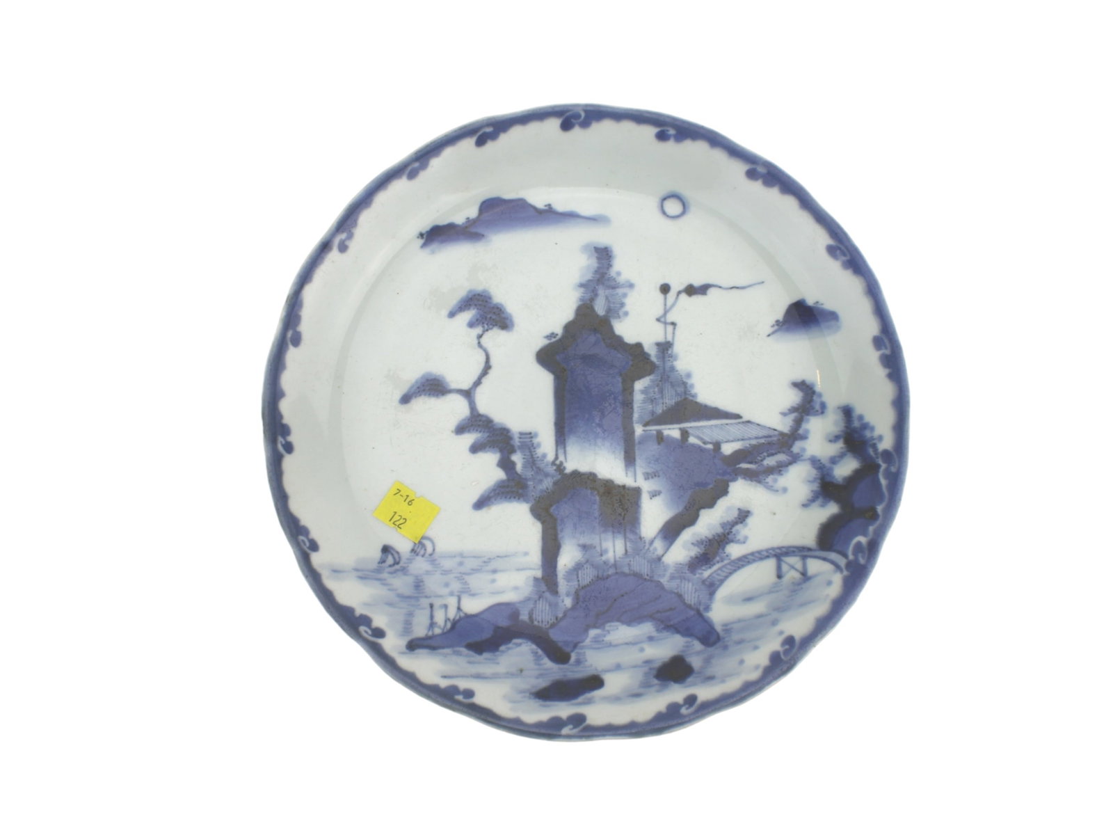 Asian Blue And White Plate: A blue and white porcelain dish with a scalloped rim, featuring a painted riverside landscape with mountains, trees, boats, a tall pavilion, and a bridge. Decorative cloud motifs adorn the border.