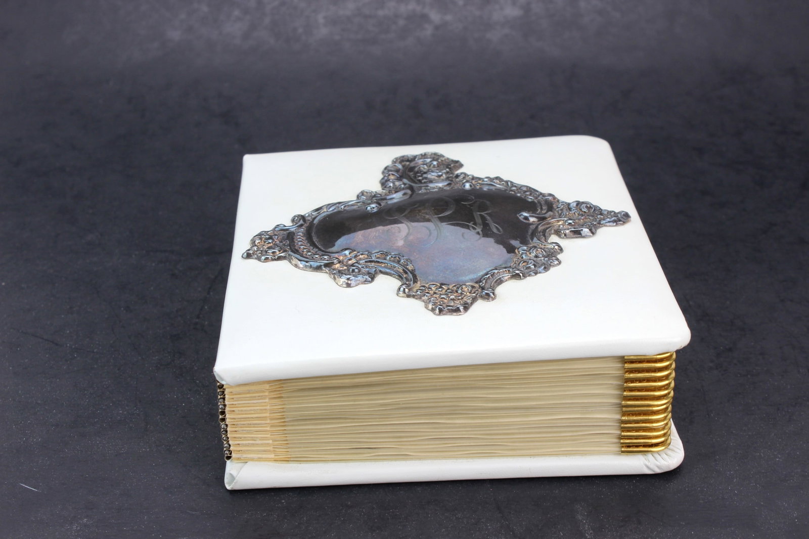 White Photo Album w Sterling Centerpiece: White photo album featuring a decorative sterling silver centerpiece with embossed monogram and gold-finished page edges. Measures approx. 5 1/2 in. (L) x 5 in. (W) x 1 1/2 in. (H). Shows light wear,