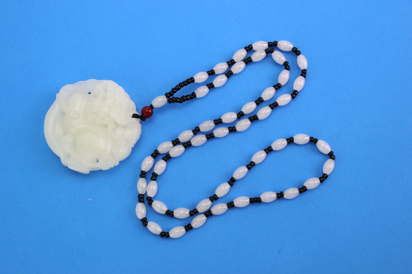 White Hardstone Pendant Necklace: Carved white hardstone pendant necklace with intricate designs, featuring a 2 in. (D) pendant on a 25 in. (L) beaded strand of white oval beads, black spacers, and a red accent bead. Well-polished