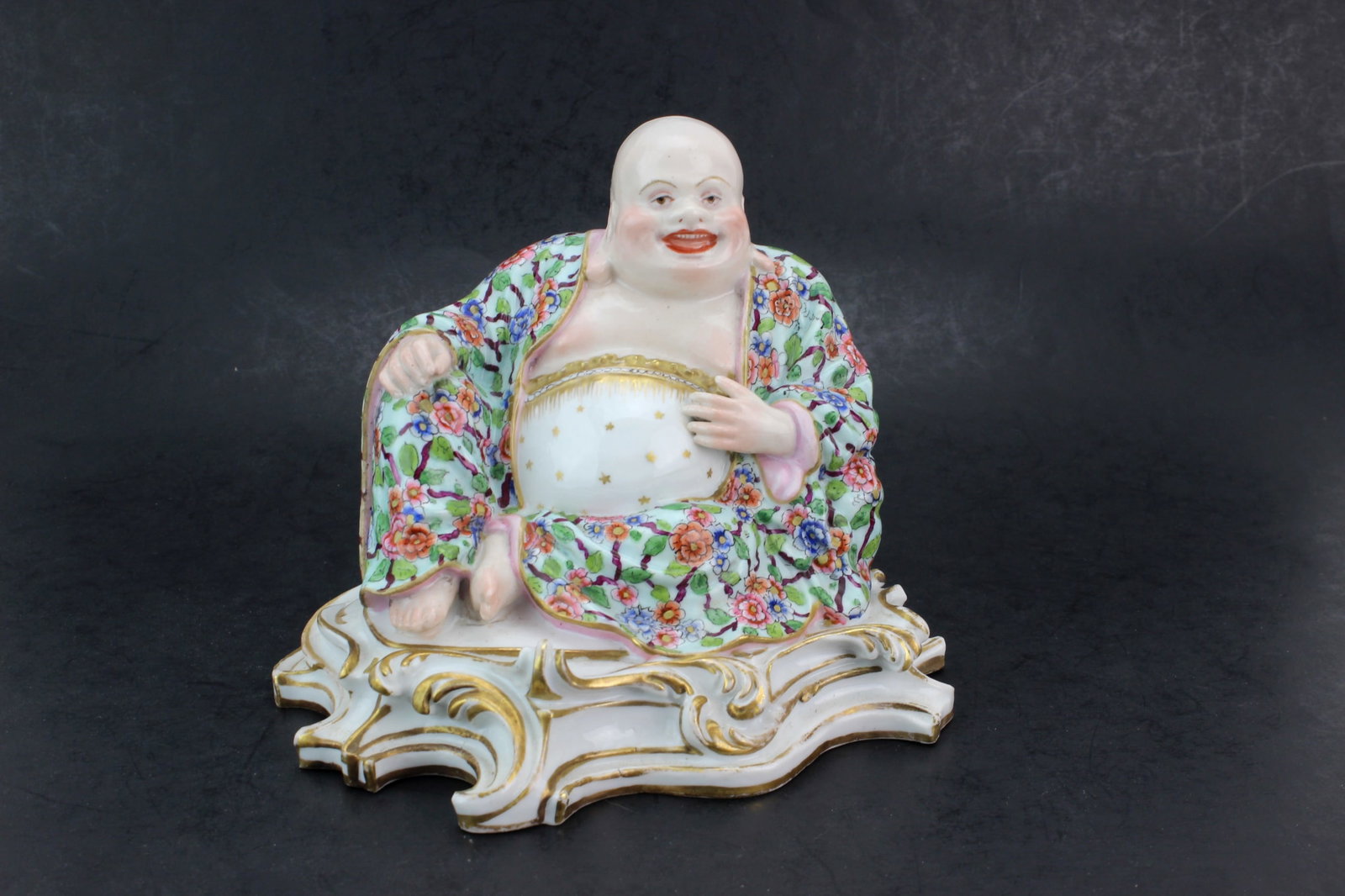 Meissen Famille Rose Porcelain Buddha: Germany Meissen porcelain laughing Buddha figure in famille rose floral robe with gilt decorated base. Measures approximately 6 in. (H) X 7 in. (L) X 6 in. (W). Appears in good overall condition with 