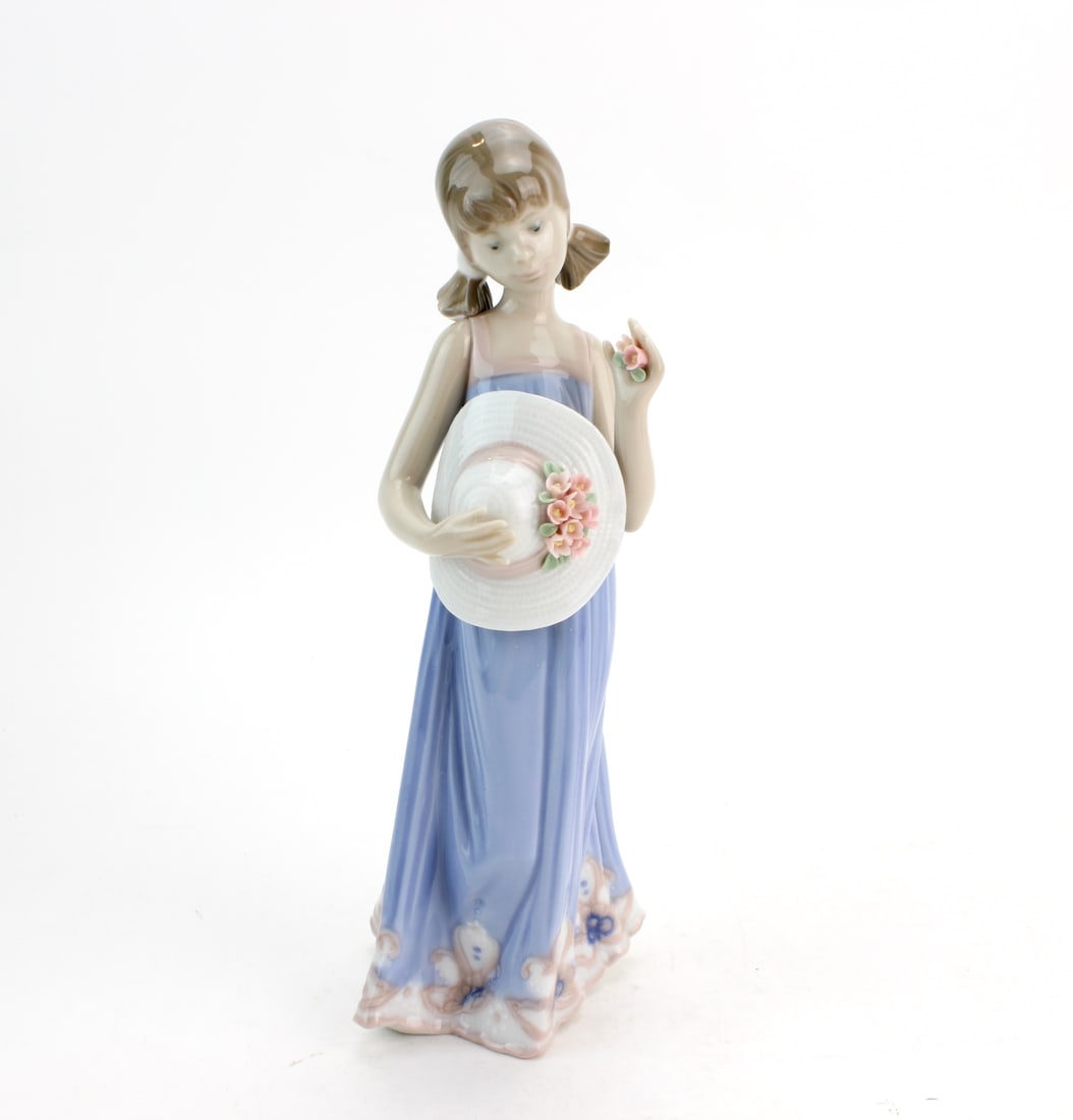 Lladro Courtney Figurine w/ Box: Lladro porcelain figurine Courtney No. 5648, depicting a young lady in a flowing blue dress holding a floral hat, complete with original box and paperwork. Figurine measures 8 in. (H); box measures 11