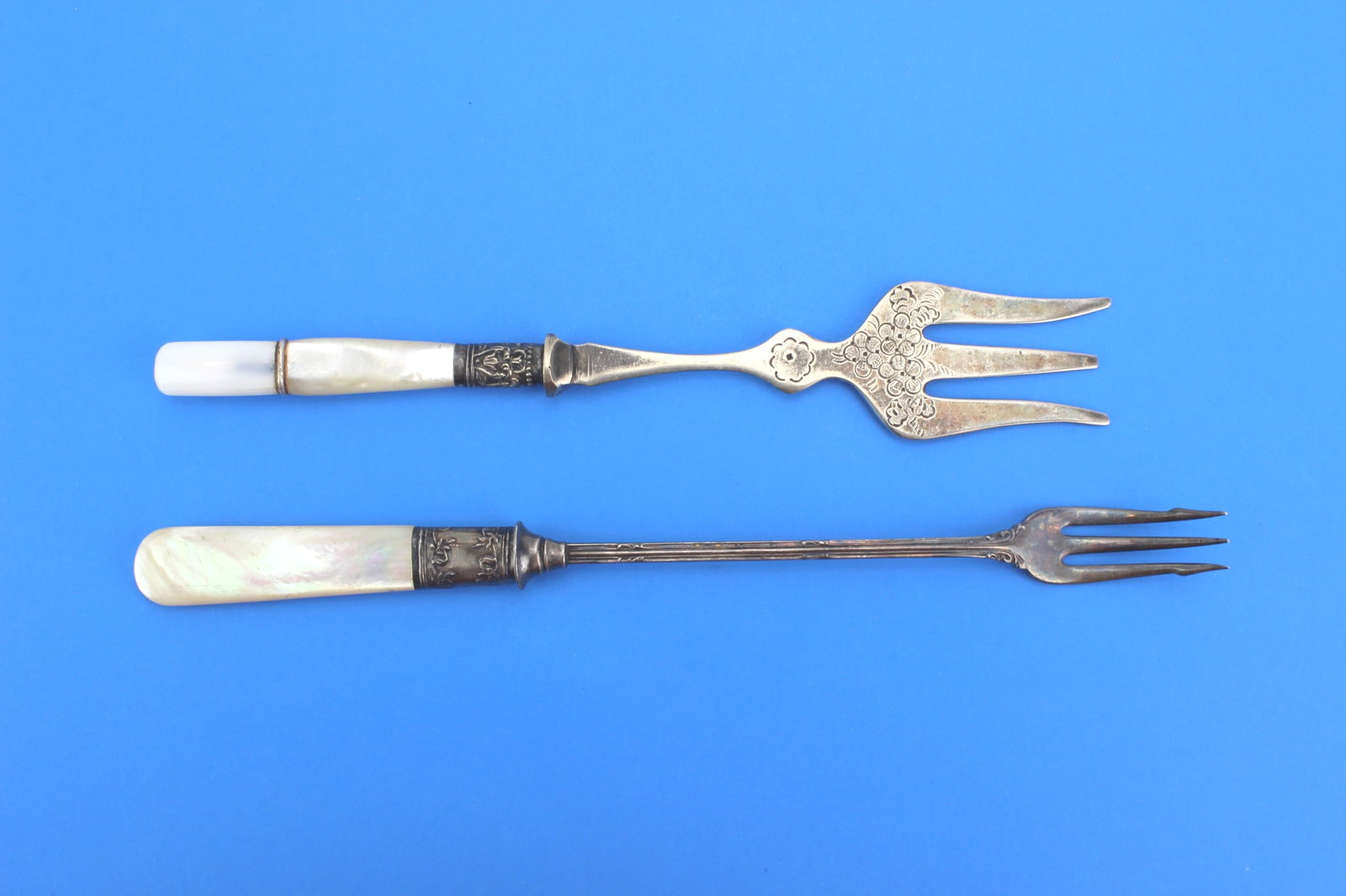 Silver Plate Forks w/ MOP Handles: Pair of silver plate serving forks with mother-of-pearl handles, including one ornate floral example. One with sterling connection. The longer fork measures 8 1/2 in. (L). Tarnish, surface wear, and