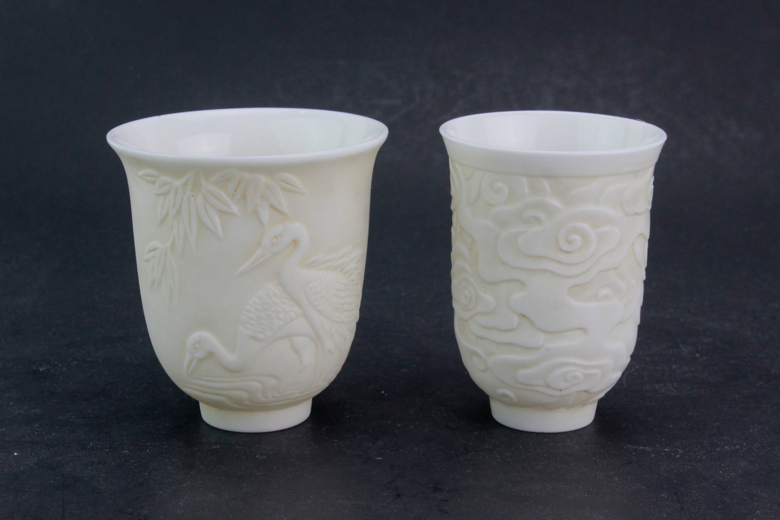 Two Chinese White Porcelain Tea Cups: A pair of blanc de chine porcelain tea cups, one decorated with cranes and bamboo, the other with cloud motifs. Finely carved in low relief, with smooth glazed interiors. Likely late Qing or Republic