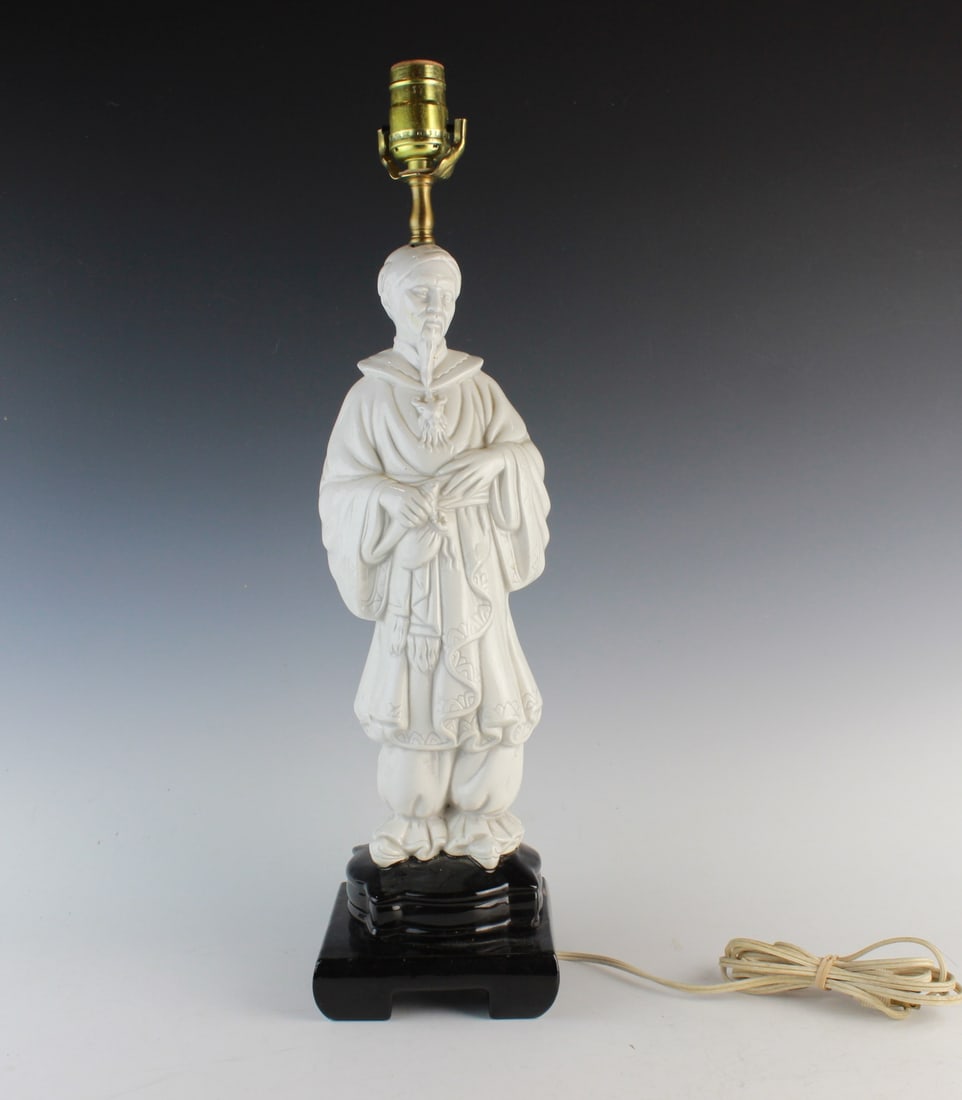 Chinese White Porcelain Figural Table Lamp: A vintage lamp with a white glazed porcelain figure in traditional Chinese dress on a black base, topped with a brass socket. Shows minor wear and small chips. The lamp measures Approx. 21 in. tall,