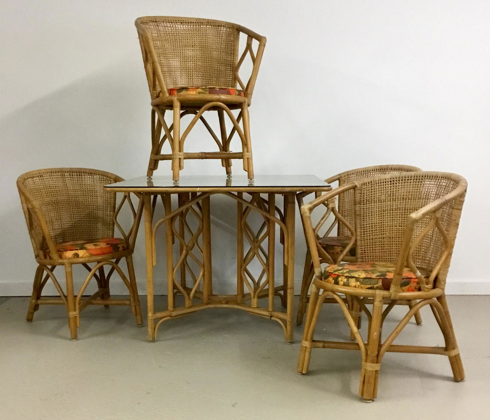 Rattan Table and Chair Set with Glass Top: Vintage bamboo and woven rattan set with four armchairs and square glass top table. Floral vinyl seat cushions. Table is 40” square and 30” tall. Each chair is 23” wide and 33”