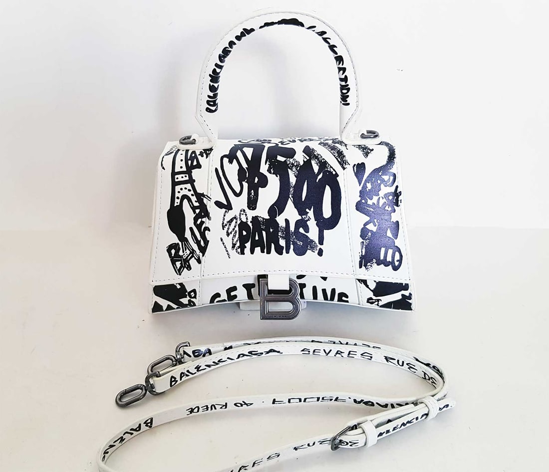 BALENCIAGA HOURGLASS XS TOTE BAG GRAFFITI: BALENCIAGA HOURGLASS XS TOTE BAG in GRAFFITI PRINTED LEATHER White leather Hourglass XS graffiti top-handle bag from Balenciaga. Black graffiti atop a white leather makes this XS Hourglass tote your