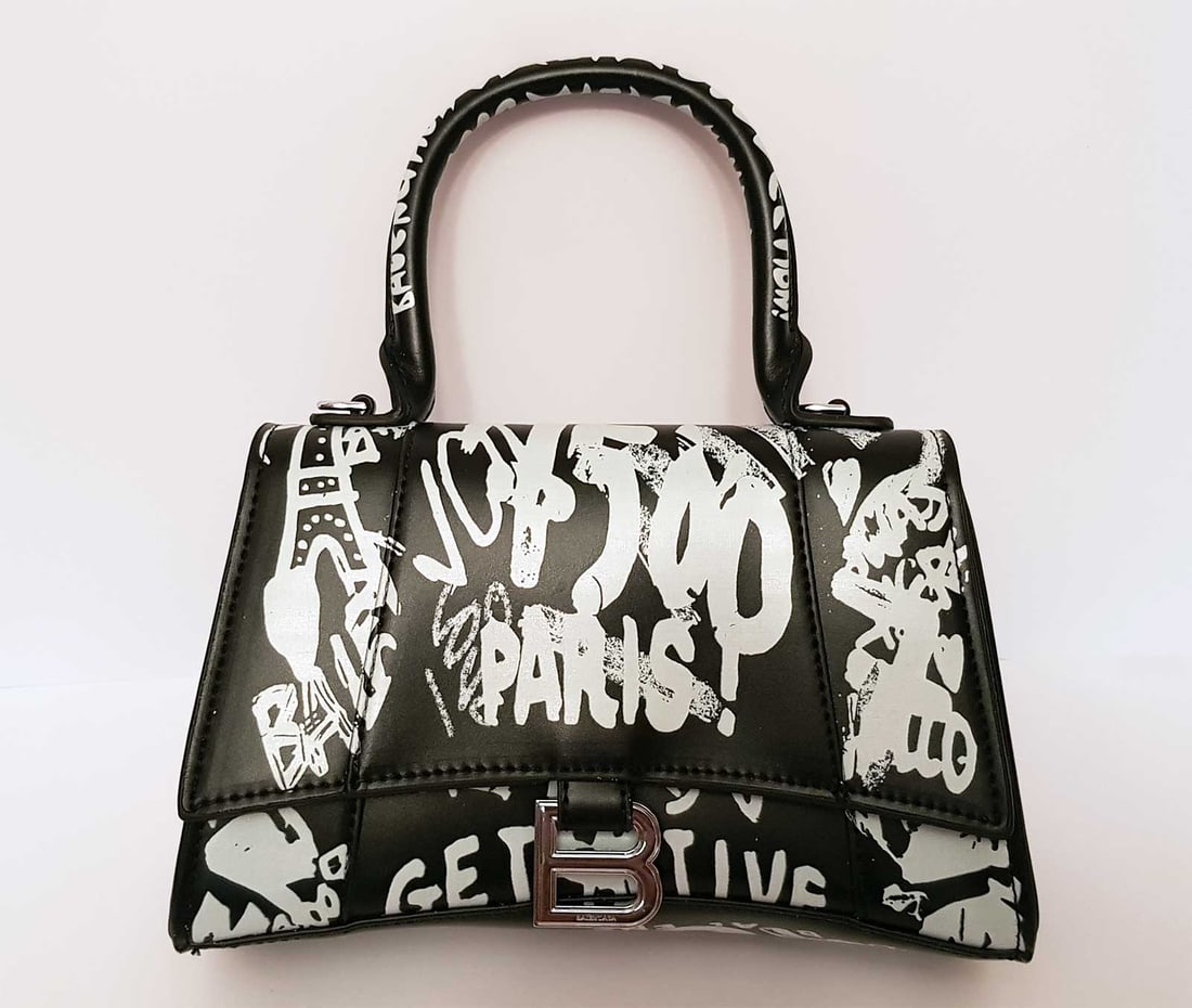 BALENCIAGA HOURGLASS XS TOTE BAG GRAFFITI: BALENCIAGA HOURGLASS XS TOTE BAG in GRAFFITI PRINTED LEATHER Black leather Hourglass XS graffiti top-handle bag from Balenciaga. White graffiti atop a black leather makes this XS Hourglass tote your n