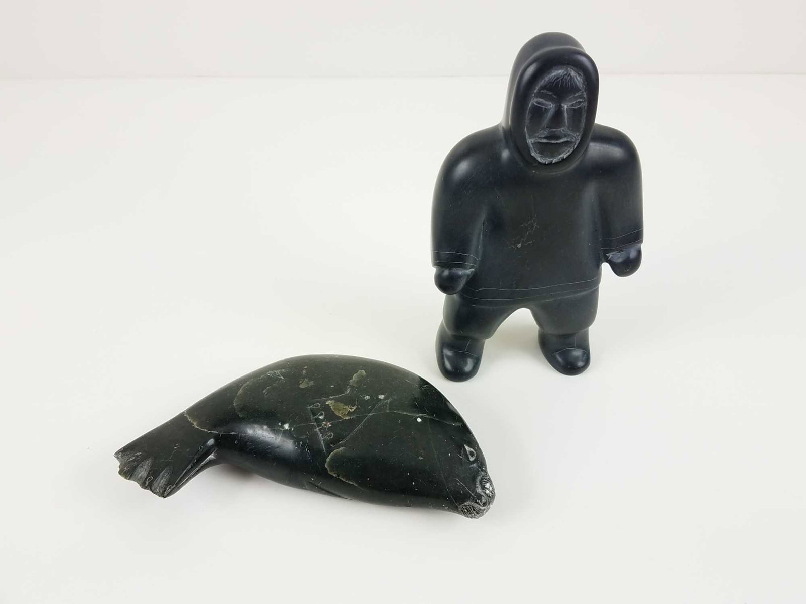 Two (2) Inuit Soapstone Sculptures: This is a lot of two (2) Inuit soapstone sculptures. Includes: One (1) sculpture of a standing Inuit hunter. Sticker on his foot reads Sarah Kudluaroq Sanikiluaq 89. MEASUREMENTS: 6" x 3.5" x 1.5" One
