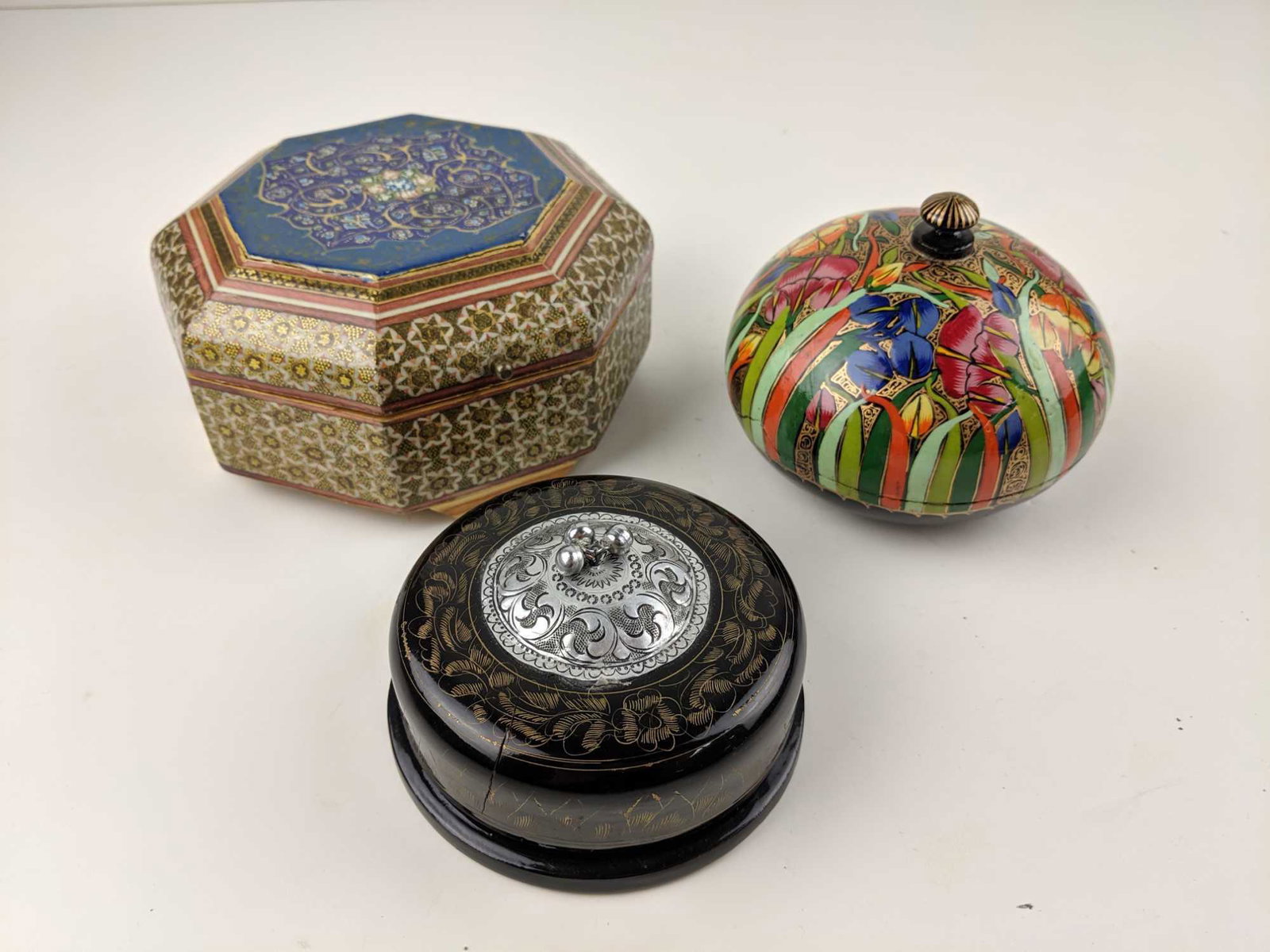 Three (3) Middle Eastern Style Trinket Boxes: This is a lot of three (3) Indian/Middle Eastern painted trinket boxes. Includes: One (1) black round box with a silver top with little bells and gold leaf design. MEASUREMENTS: 4" diameter, 1.5" tall