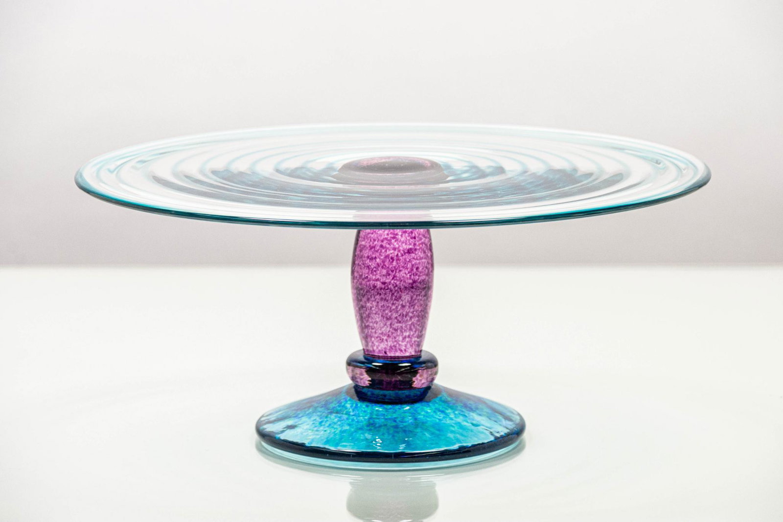 Title Art Glass Cake Stand: DescriptionClear art glass cake stand with blue and magenta accents. CONDITION: Excellent DIMENSIONS: H 5” x W 11.5" x D 11.5"