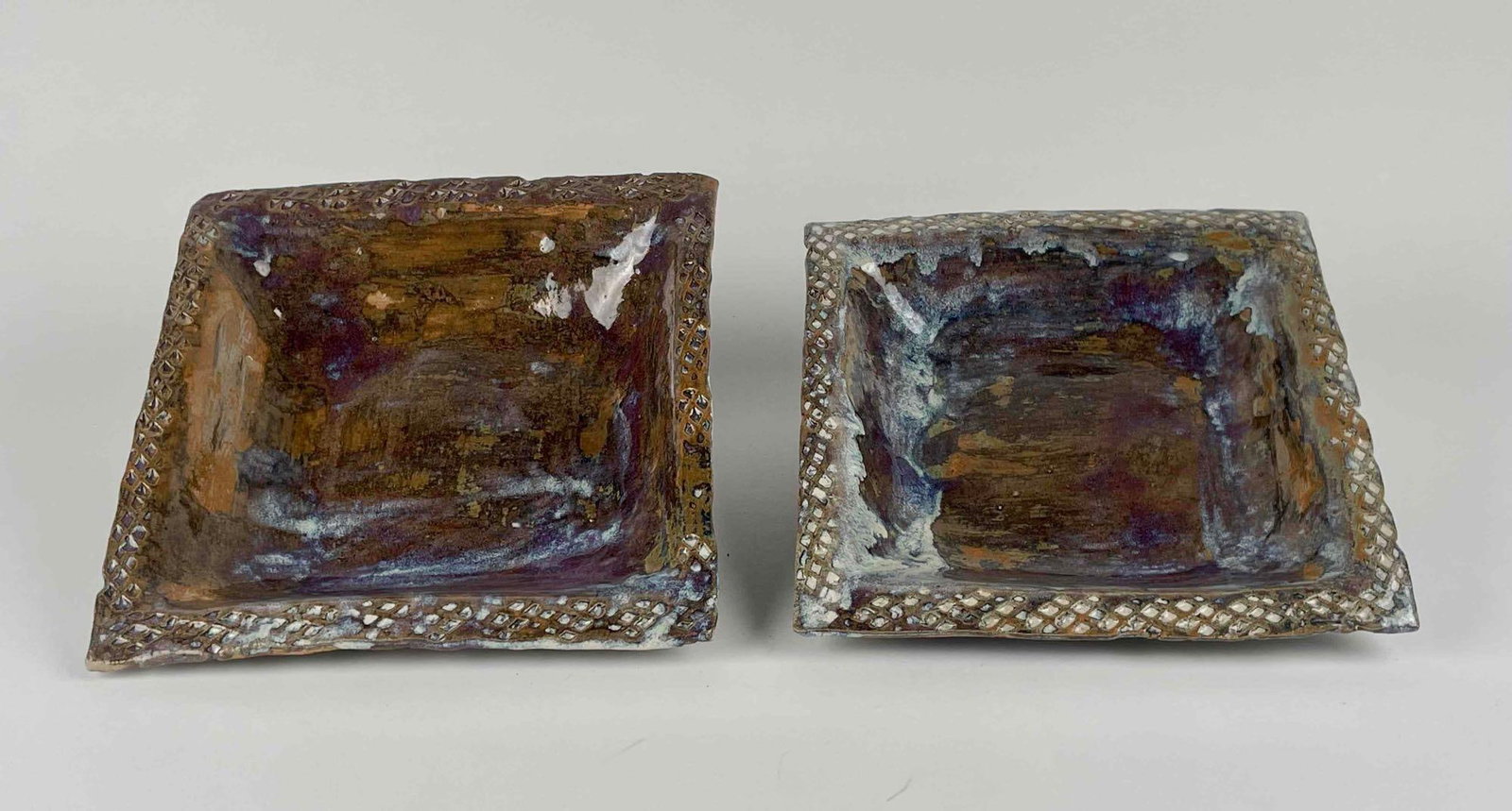 Pair of Handmade Shallow Square Bowls: Pair of handmade shallow square slab worked bowls. Glazed colors include earth tones with light blue, and lavender. Bowls have a basket weave pattern around edges. Artist signature is present on the b