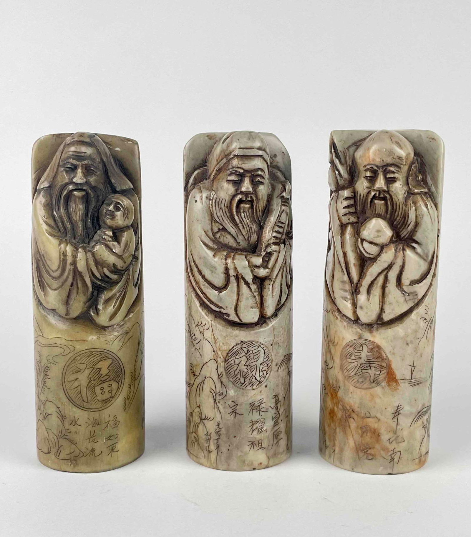 Three Chinese Soapstone Carved Sealing Stamps: Three Chinese carved soapstone seals, 20th century, each of cylindrical shape, relief carved with a god or sage above incised text, with an incised landscape view to each verso. From the home of Jon J