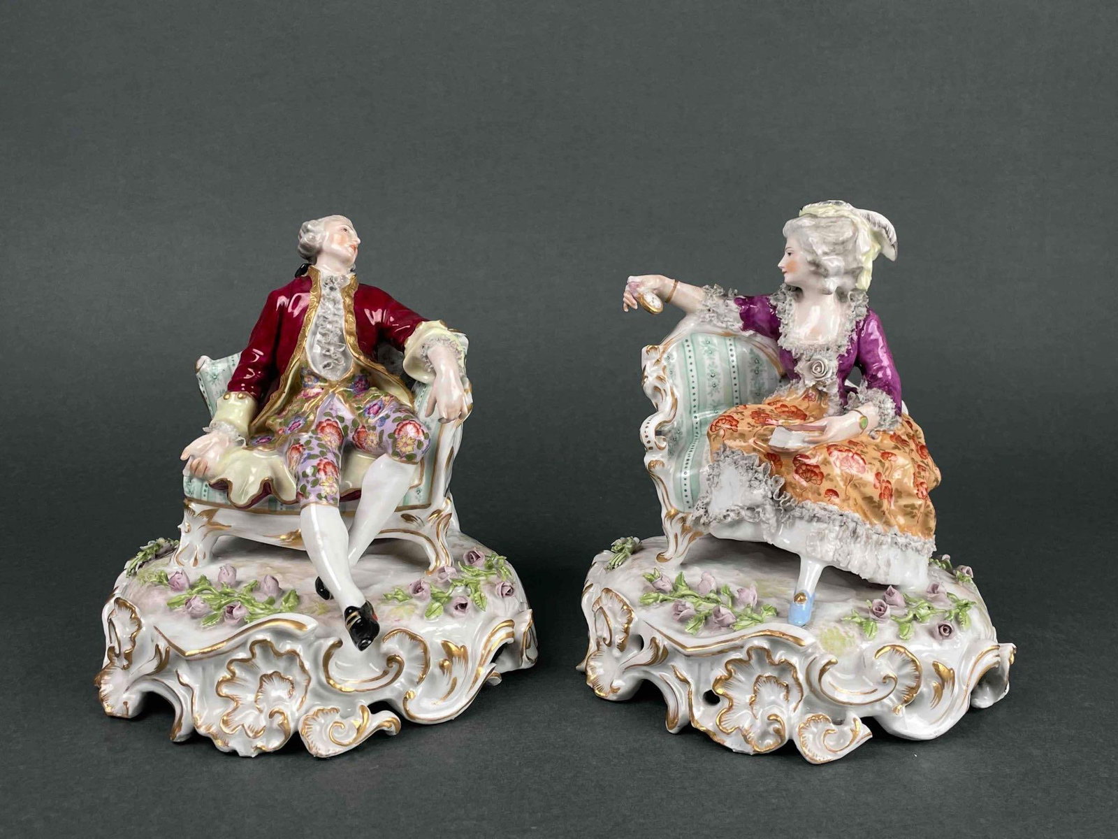 Pair of Meissen Porcelain Figurines (18th Century): Pair of Meissen porcelain figurines. Man and woman sitting on separate chairs. Woman checks her pocket watch as man looks to his side. Base is decorated with gilted design and floral detailing. In goo