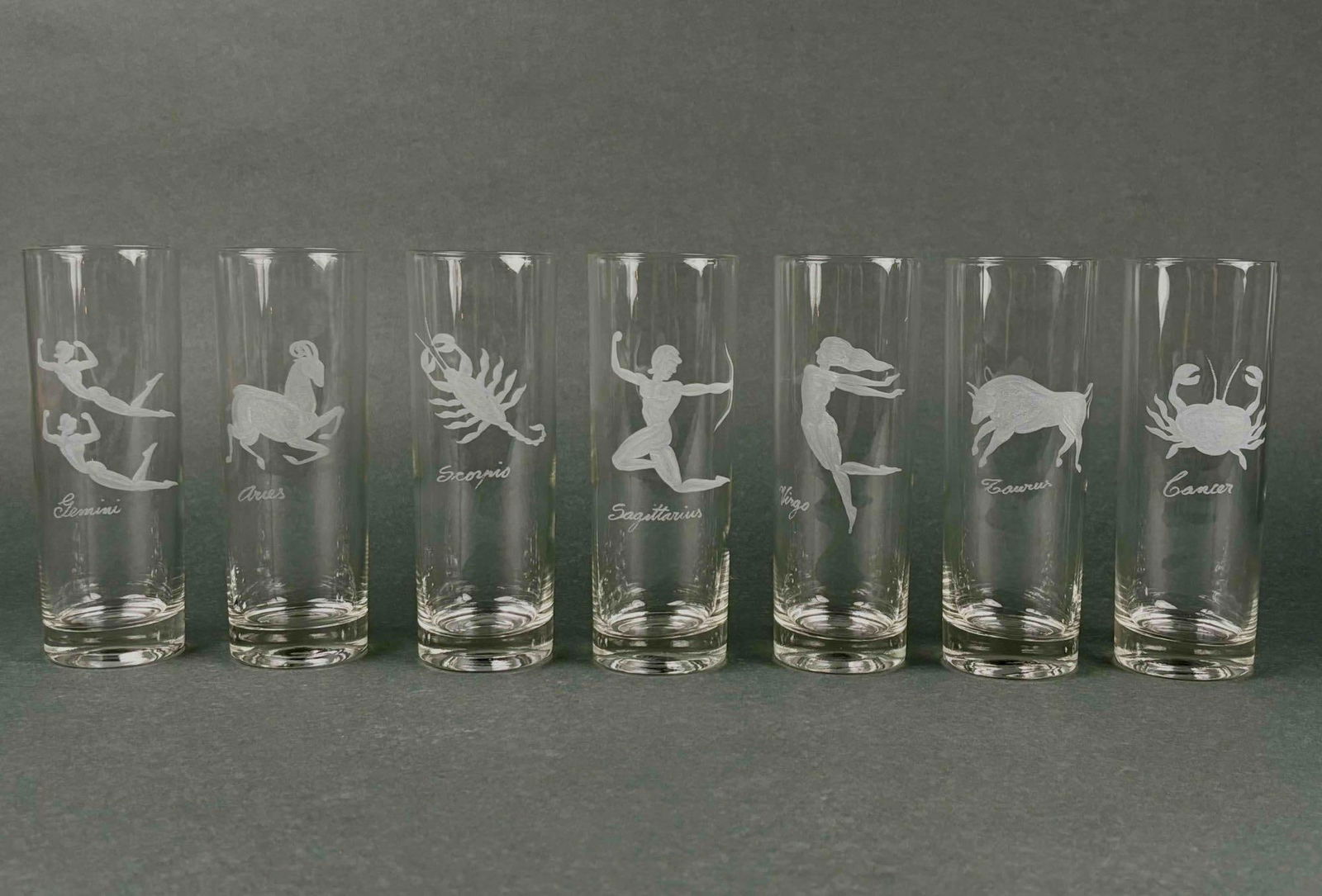 7 Etched Zodiac Collins Glasses: 7 Tom Collins glasses depicting zodiac symbols in etched glass. All glasses are in excellent condition with little to no signs of wear or use. Zodiac is incomplete. Signs represented are Cancer, Gemin