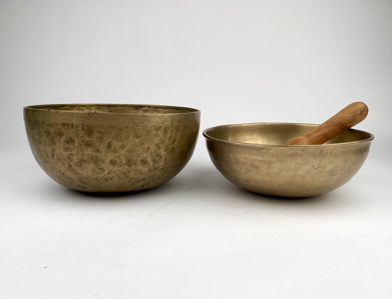 Two Brass Handmade Tibetan Singing Bowls with Wooden Striker: A set of two brass singing bowls, each showcasing unique hand-crafted textured surfaces. Includes a wooden striker. Condition shows signs of wear consistent with age and use, including surface marks a