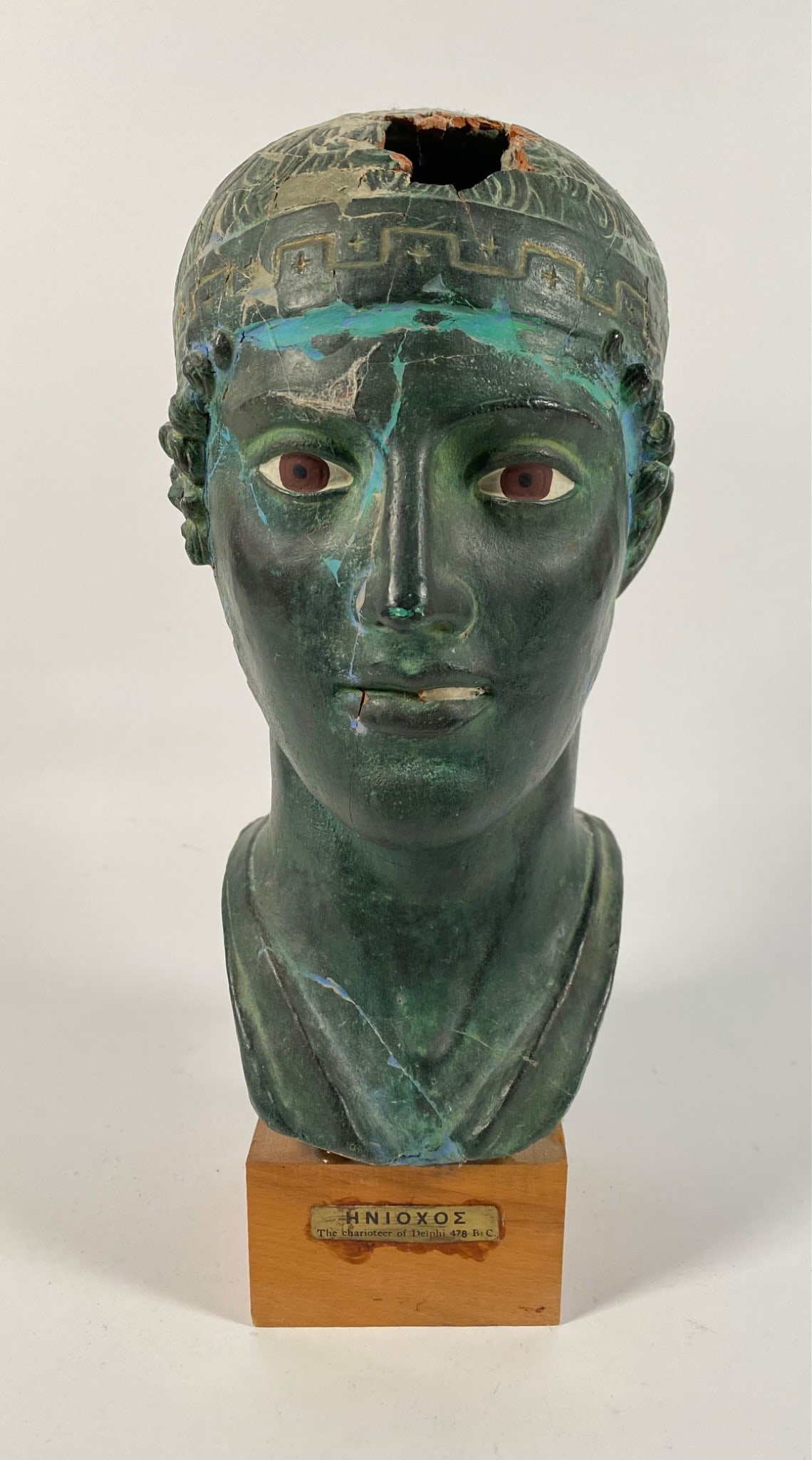 Bronze Bust of Charioteer of Delphi on Wood Base: Bronze bust replica representing â€˜The Charioteer of Delphiâ€™, mounted on a wooden base. Features include a detailed headdress and facial features, with evident pati