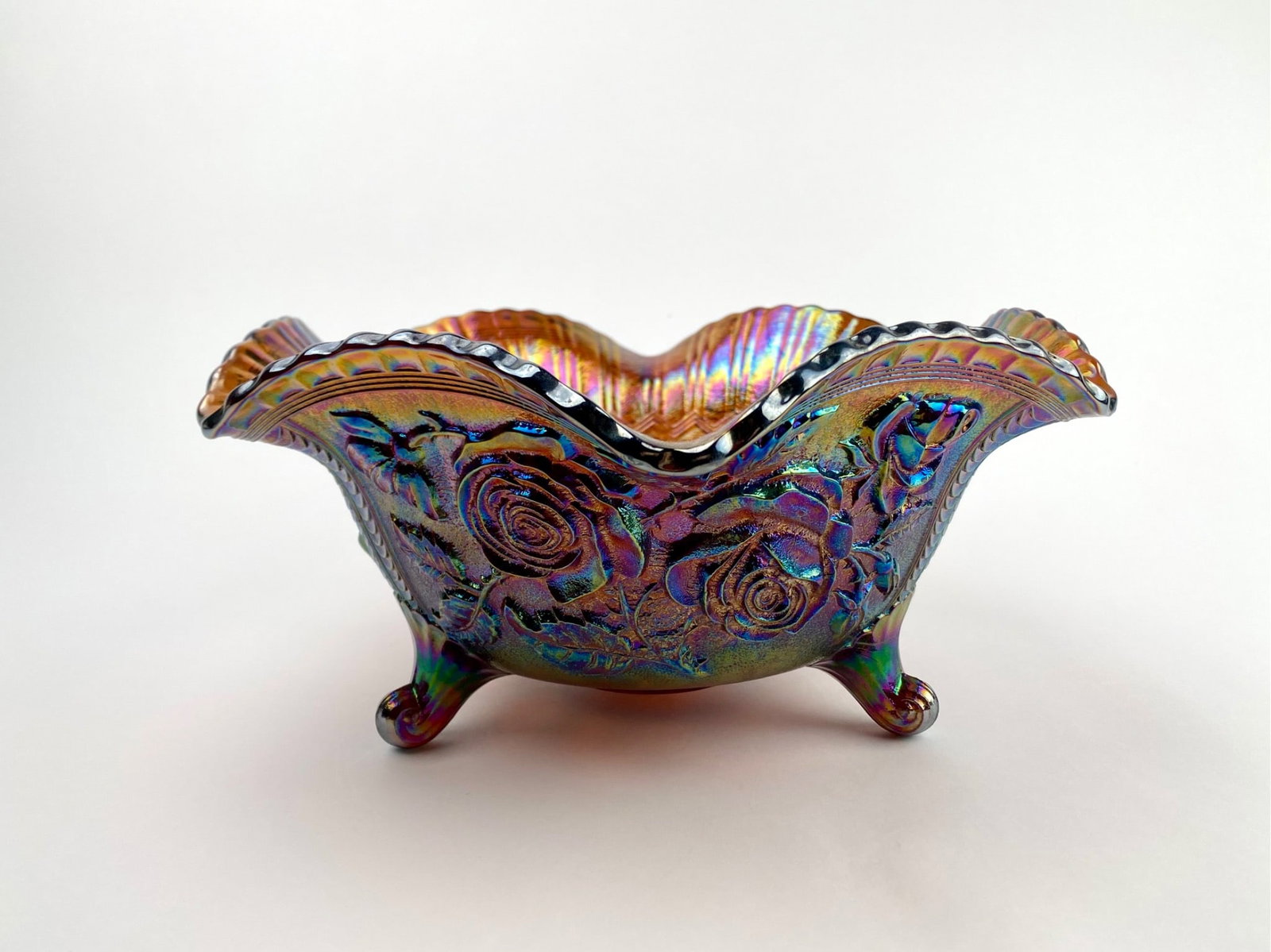 Imperial Carnival Glass Footed Bowl: An iridescent carnival glass bowl featuring an intricate rose pattern on the exterior and interior. The bowl has a scalloped edge and stands on four short feet. The iridescent finish displays a range 