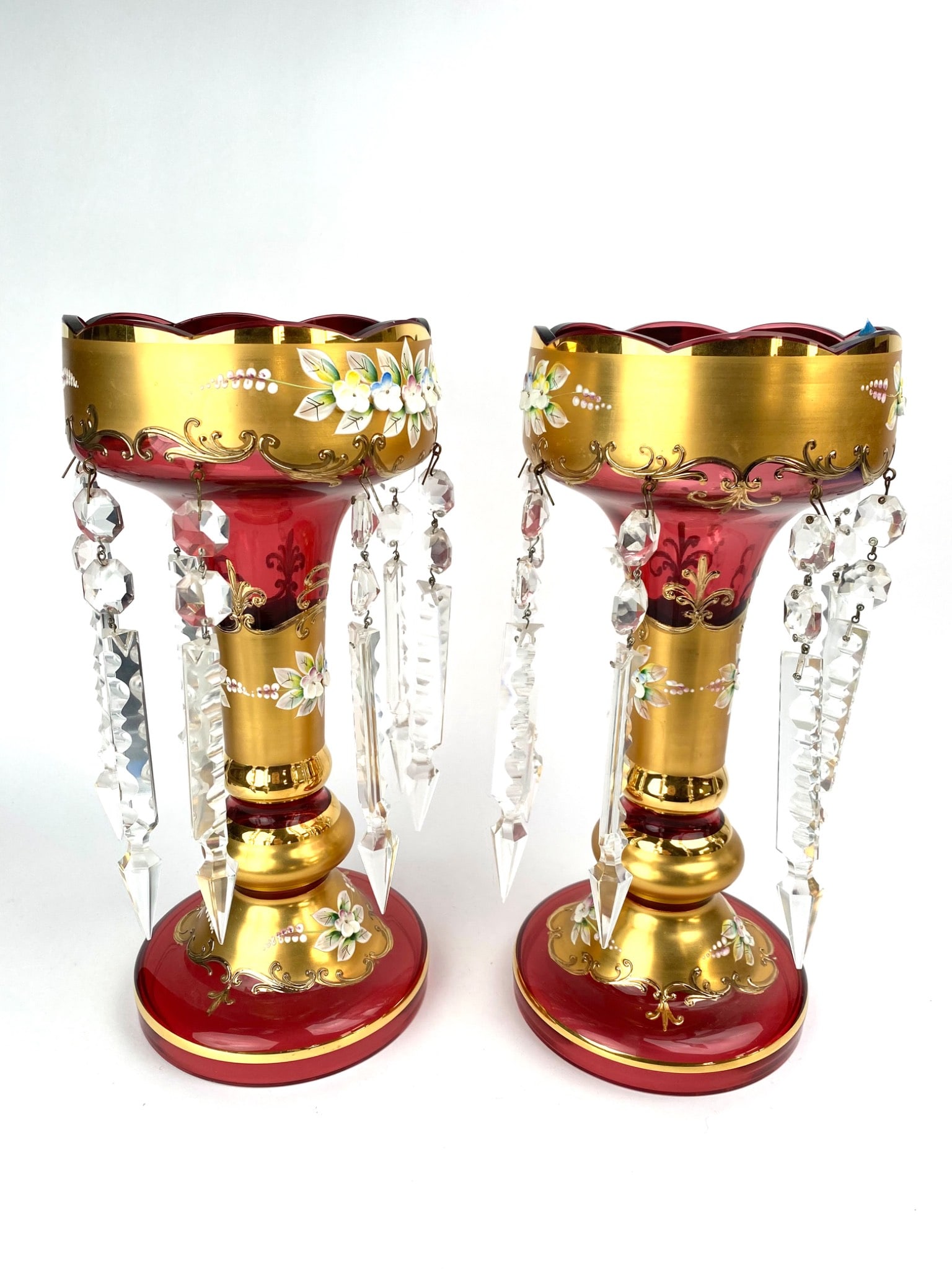 Victorian Bohemian Glass Mantle Lusters: Pair of Bohemian cranberry glass mantle lusters featuring a gilt base with applied hand-painted floral designs. Each luster is adorned with hanging glass prisms. The scalloped rims and vibrant cranber