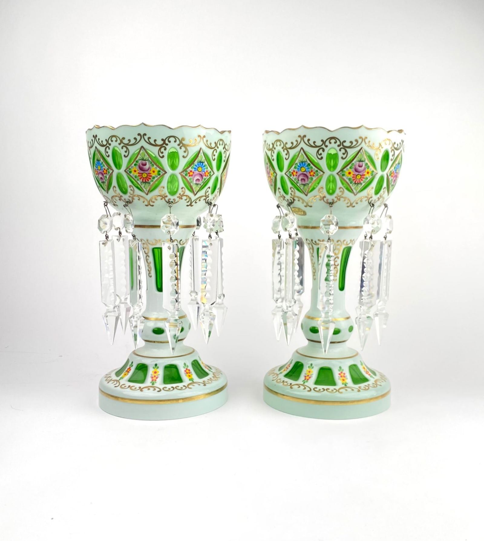 Pair of Bohemian Glass Mantle Lusters: Pair of vintage Bohemian glass mantle lusters, featuring green overlay with detailed floral and gilt decoration. Each luster includes hanging faceted crystal prisms and scalloped edges. Intricate enam