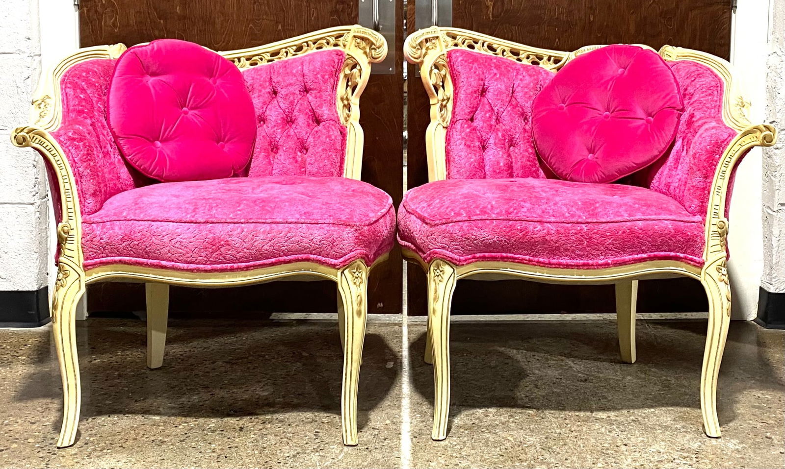 Pair of Carved Armchairs with Pink Upholstery and Button-Tufted Cushions: Pair of intricately carved armchairs featuring cream-painted wooden frames with floral and scroll motifs, complemented by vibrant pink fabric upholstery. The chairs include button-tufted cushions and