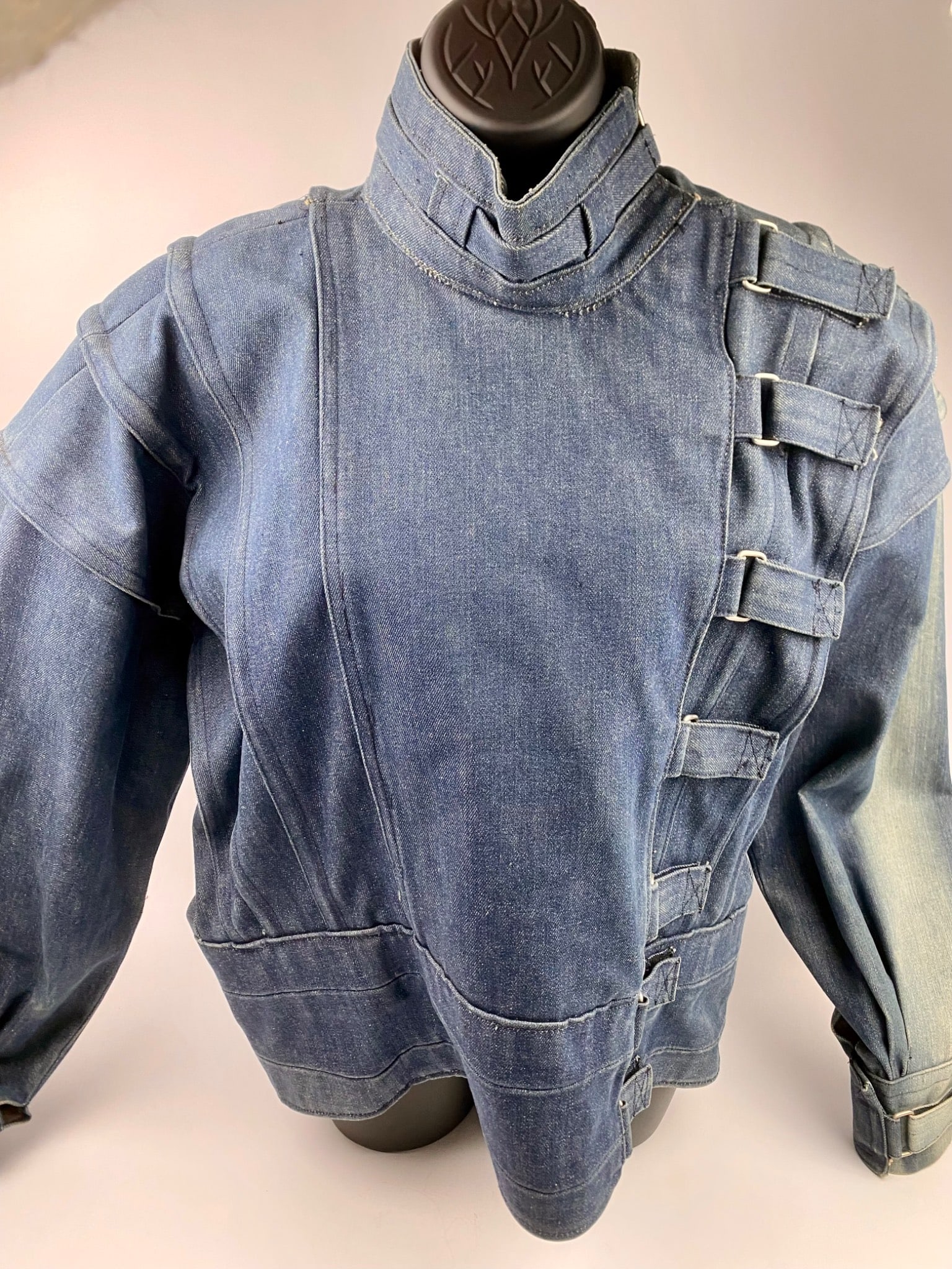 Denim Jacket By Wheeler With Buckle Accents: Denim jacket designed by Tony Gomez for By Wheeler, featuring multiple buckle accents along the front closure and a high neck collar with additional buckle detailing. The jacket includes unique detail