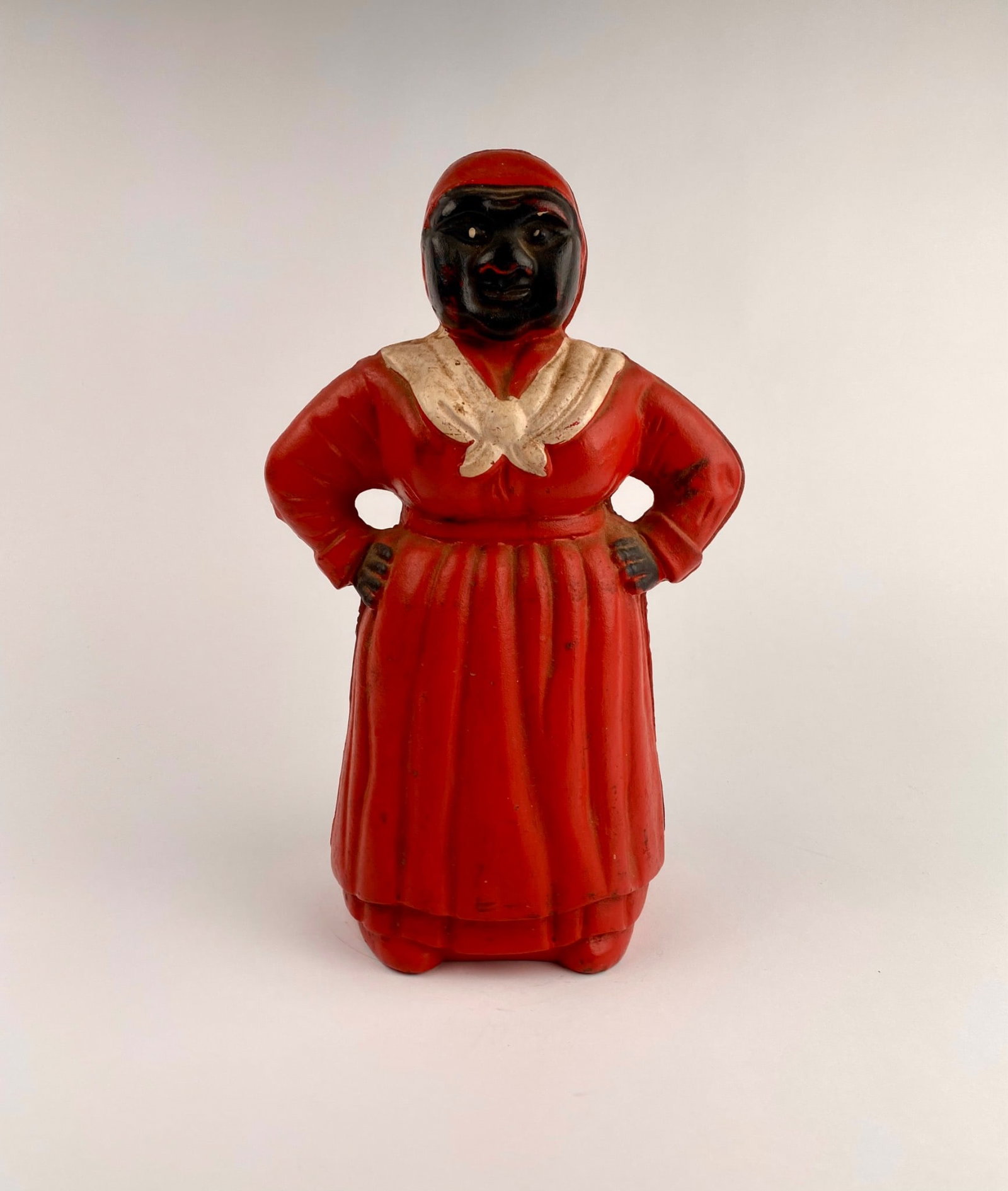 Cast Iron Aunt Jemima Coin Bank: Cast iron coin bank depicting a Aunt Jemima wearing a red dress and white shawl. Features a coin slot at the back of the figure. The figure is adorned with hand-painted accents, including facial detai