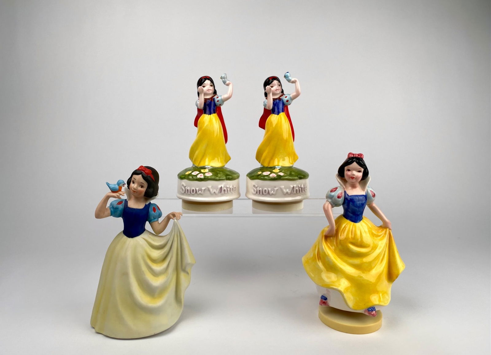 Set of Four Ceramic Snow White Music Boxes: Set of four painted ceramic Snow White musical figurines. The music boxes feature Snow White holding a bird, labeled Snow White on circular bases. The other two figures depict Snow White in distinct p