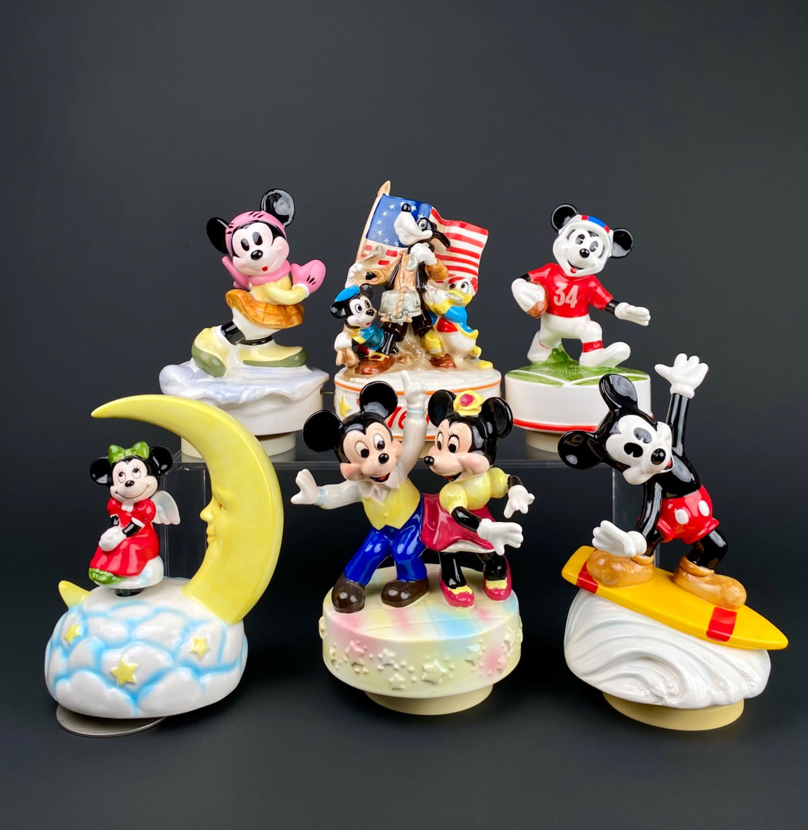 Set of 7 Disney Mickey and Minnie Music Boxes: Set of 7 collectible Disney music boxes depicting Mickey and Minnie Mouse in various themed designs, including surfing, moon scenes, 1776 to 1976 patriotic representation, and others. Each figurine is