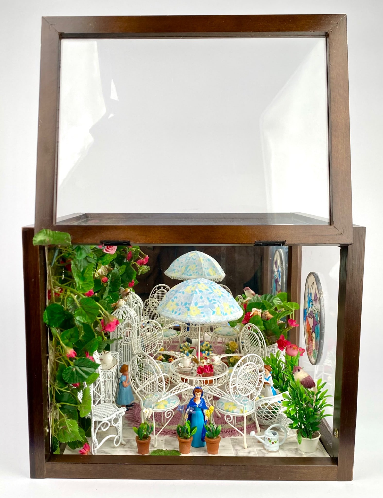 Glass Enclosed Diorama Disney Garden Scene: A glass enclosed miniature garden scene. The diorama features intricate white wire furniture, a floral patterned umbrella, potted plants, Disney figures and characters, and detailed decor, all set wit