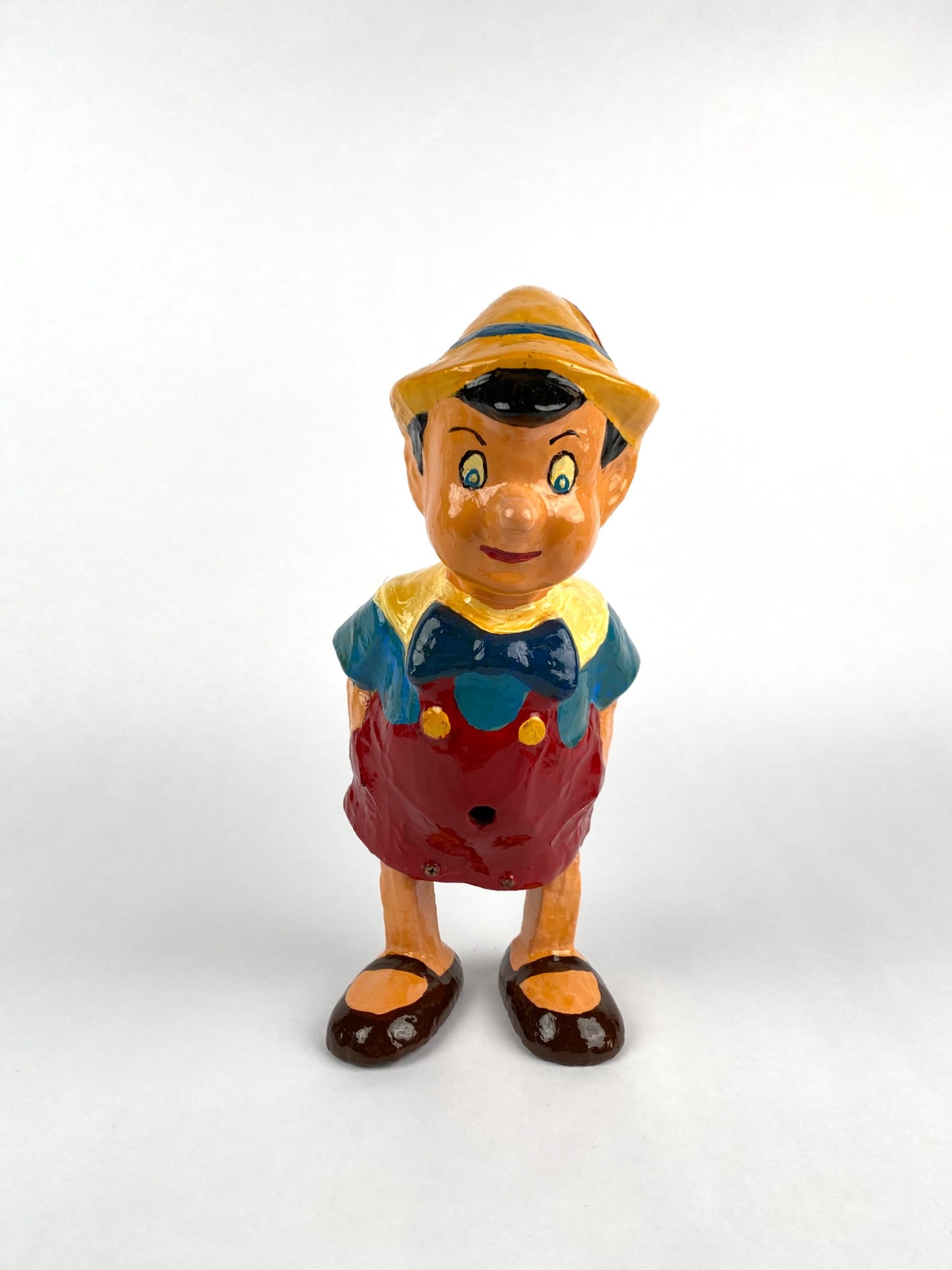 Hand-Painted 1940s Pinocchio Figure from France: Hand-painted 1940s Pinocchio figure. Made of painted metal, featuring vibrant colors including red, yellow, blue, and brown. The figure includes a functional slot in the back for coin insertion and a 