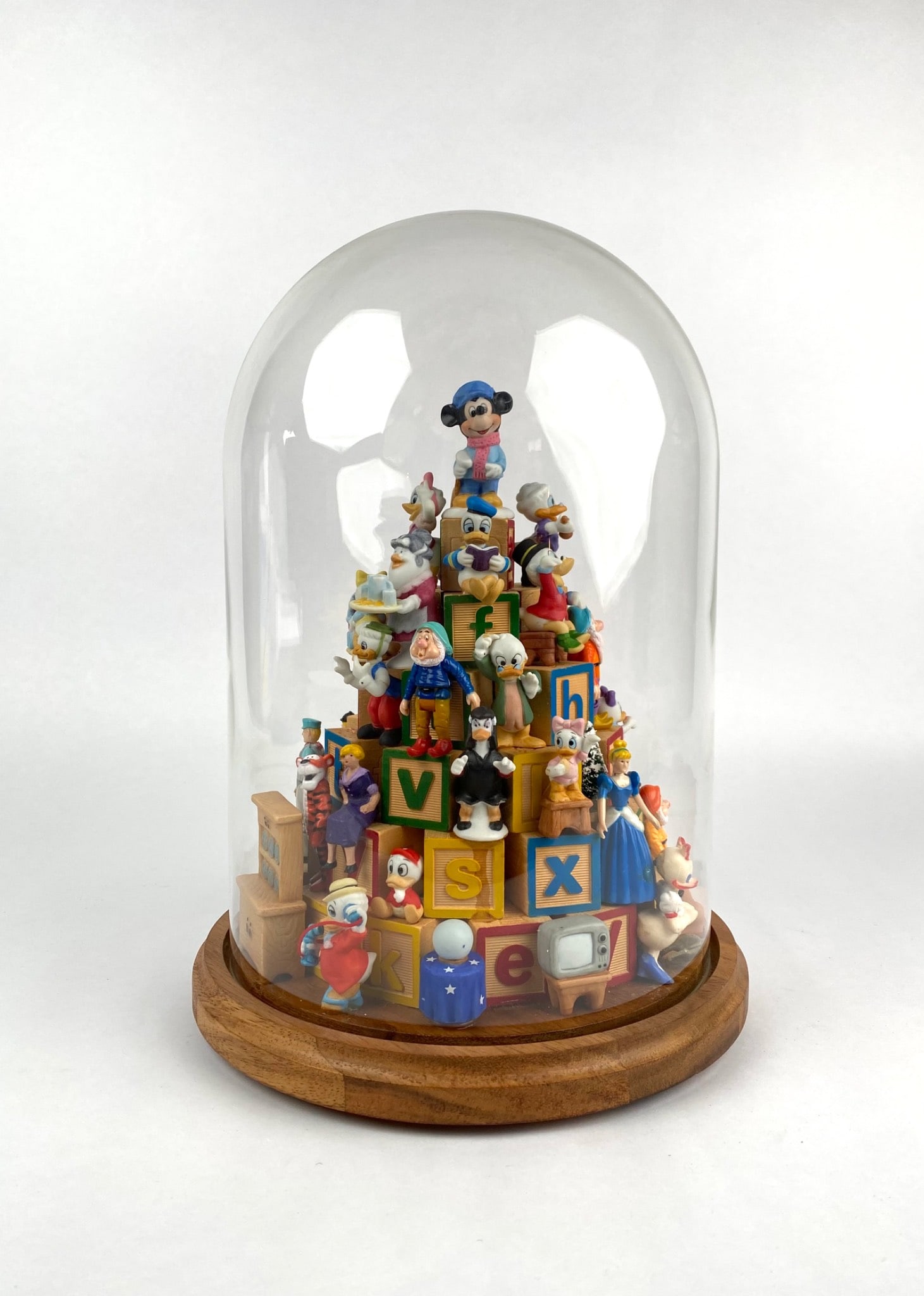 Disney Themed Diorama Featuring Figurines and Wooden Blocks: Disney themed diorama under a glass dome with a wooden base, showcasing an assortment of miniature figurines representing famous Disney characters, stacked and positioned along wooden alphabet blocks.